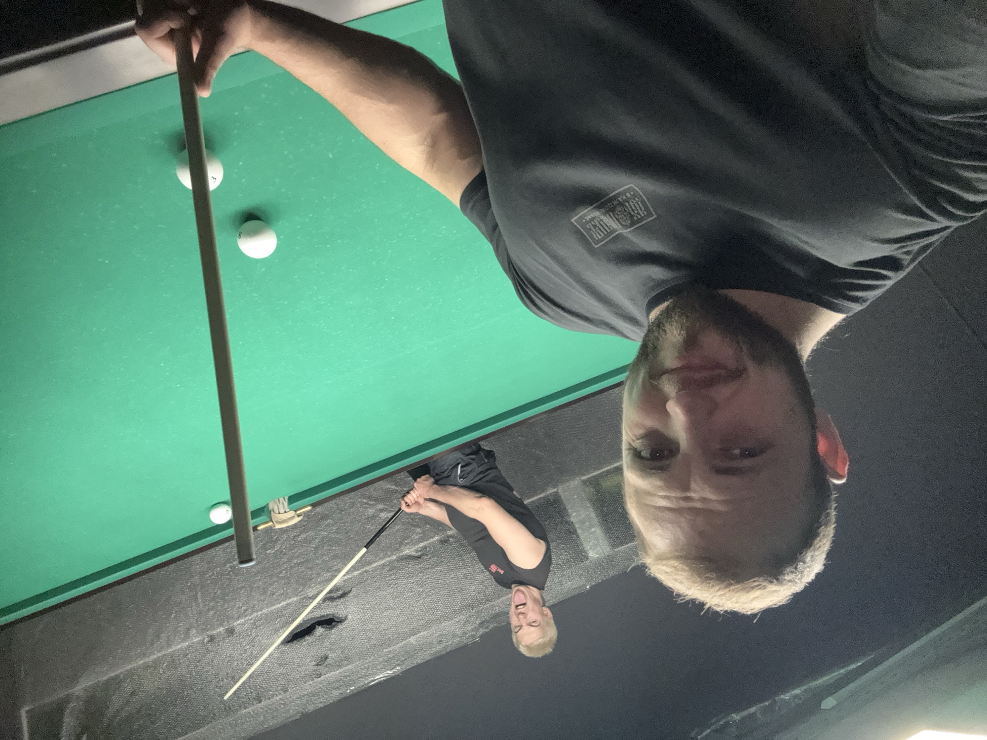 Two men are in a room with a pool table. The man in the foreground is closer to the camera and is looking directly at it. He has light brown hair, a beard, and is wearing a black t-shirt. The man in the background is further away and is holding a pool cue. He has blonde hair and is sticking his tongue out. He is wearing a black sleeveless t-shirt and shorts. The pool table is green and there are several pool balls on it. The room appears to be dimly lit.