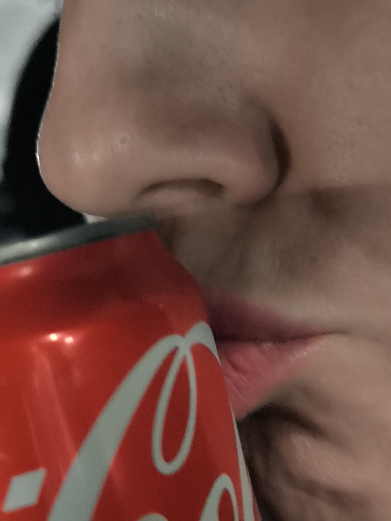 A close-up image shows a person holding a red Coca-Cola can to their lips. The nose and lower face of the individual are visible. The background is blurry and indiscernible. The location of the scene cannot be determined from the visual information provided.