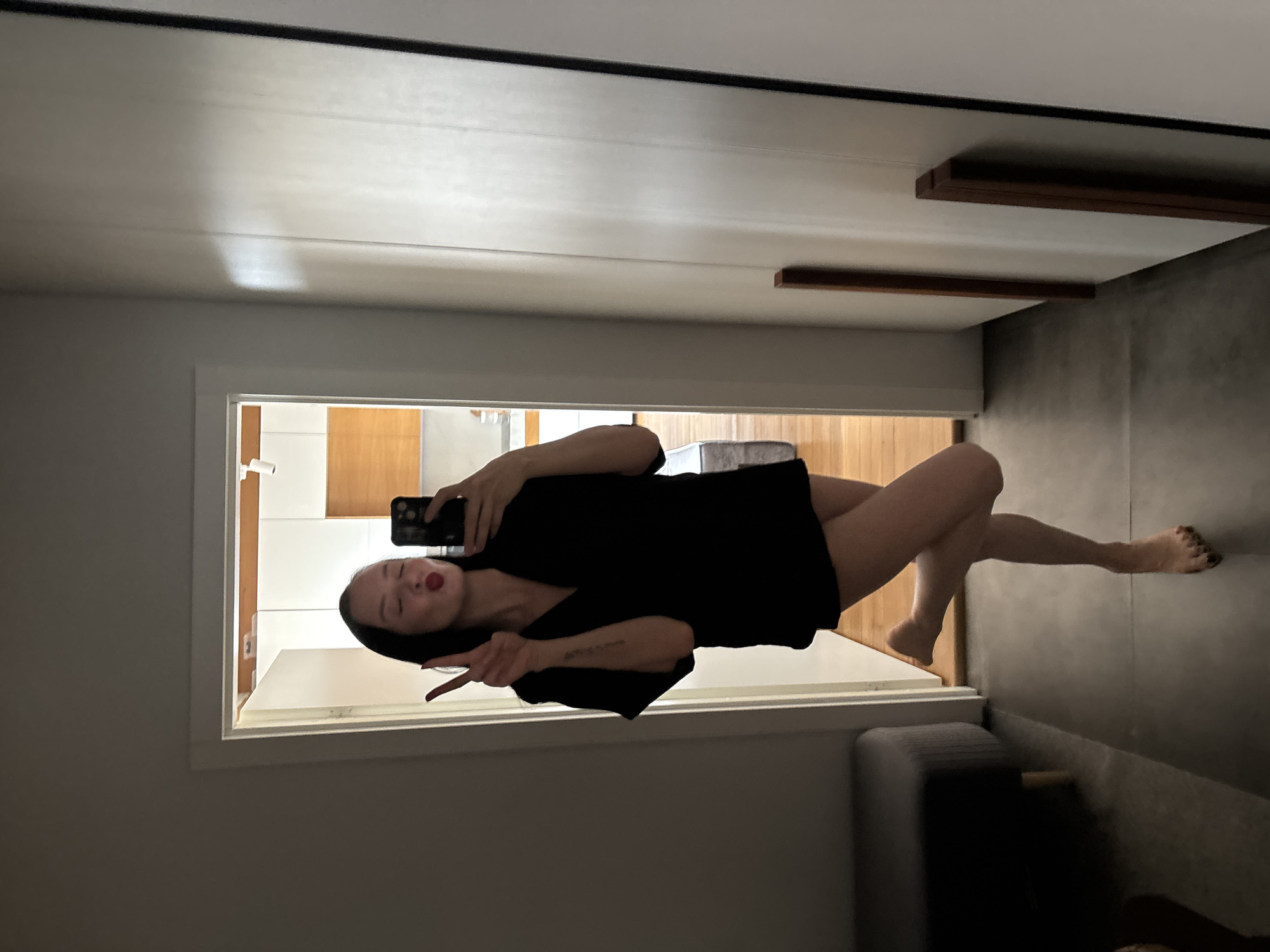 A person is captured in a mirror selfie, standing indoors. The subject, a woman with dark hair and red lipstick, is reflected pouting her lips playfully and making a peace sign with her left hand, while holding a black smartphone with her right hand to take the photo. She is wearing a loose-fitting black top or dress, with her bare legs visible. A faint, unreadable text or tattoo is visible on her left forearm.
The reflection in the mirror reveals a brightly lit room behind her, featuring white walls or cabinetry and wooden elements, possibly a kitchen or hallway. A small white object, which could be a camera or light fixture, is visible in this reflected space. In the immediate foreground of the image, a dark grey sofa or bench is partially visible. The room where the photo is taken has light grey walls and a prominent white door frame. The ceiling displays distinct white panels with a dark trim and features two decorative horizontal wooden beams. The overall lighting suggests an indoor setting, possibly during the day with indirect light. There are no specific visual cues within the image to confirm the location is Chaikovsky, Russia.