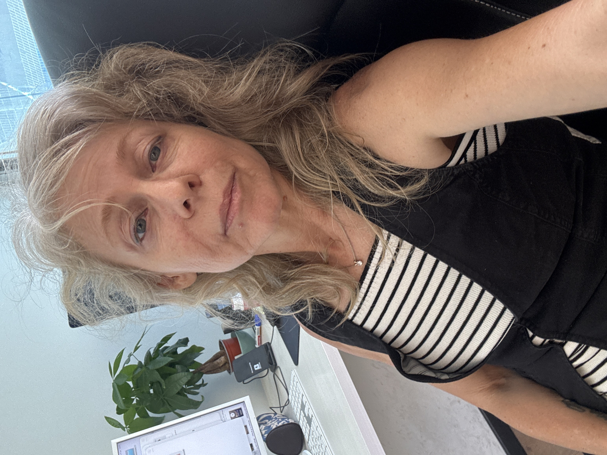 A woman is captured in a selfie-style photograph, looking directly at the camera with a calm, neutral expression. She has long, wavy hair, a mix of blonde and grey, and light-colored eyes. She is wearing a horizontally striped white and black top underneath a dark, sleeveless garment, possibly a pinafore or jumpsuit. A delicate silver necklace with a small pendant is visible around her neck.
The setting appears to be indoors, likely a home office or personal workspace, during the daytime. She is seated, with a black office chair partially visible behind her. To her left, a light blue wall forms the background, and through a window, the blurry silhouettes of tall, modern buildings can be seen, indicative of an urban environment such as Tel-Aviv, Israel.
On the white desk in front of her, several objects are arranged. These include a computer monitor displaying a graphical user interface, a white keyboard, and a patterned mouse. A small green potted plant, possibly a money tree, rests in a reddish-brown pot. Next to the plant, a black charging device with an "i" icon within a square is visible. The lighting is bright and natural, suggesting good daylight conditions.