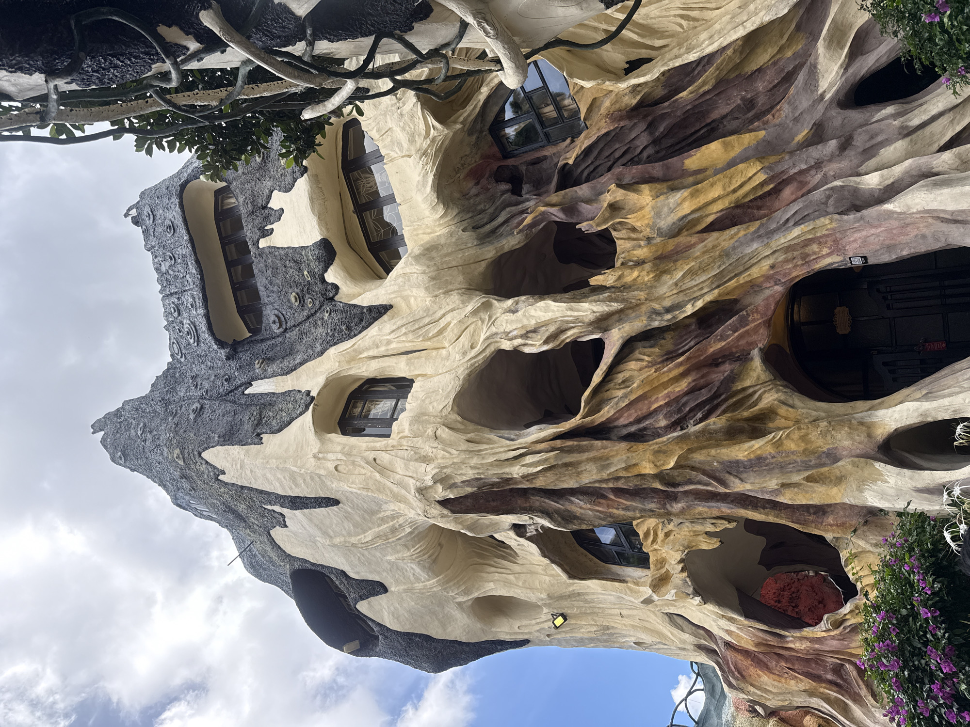 The image captures an exterior view of the Hang Nga Guesthouse, also known as the "Crazy House," in Da Lat, Vietnam. The building's facade is designed to resemble a giant, gnarled tree trunk, with organic, flowing shapes and textured surfaces in shades of cream, brown, and dark gray. Numerous irregularly shaped openings and windows punctuate the structure, some appearing as hollows or caves. The upper section of the building features a jagged, dark gray roof that mimics rock or bark, adorned with small, circular protrusions. Lush green foliage and bright purple flowers are visible at the base of the structure, contrasting with the building's unusual architecture. The sky above is partly cloudy.
