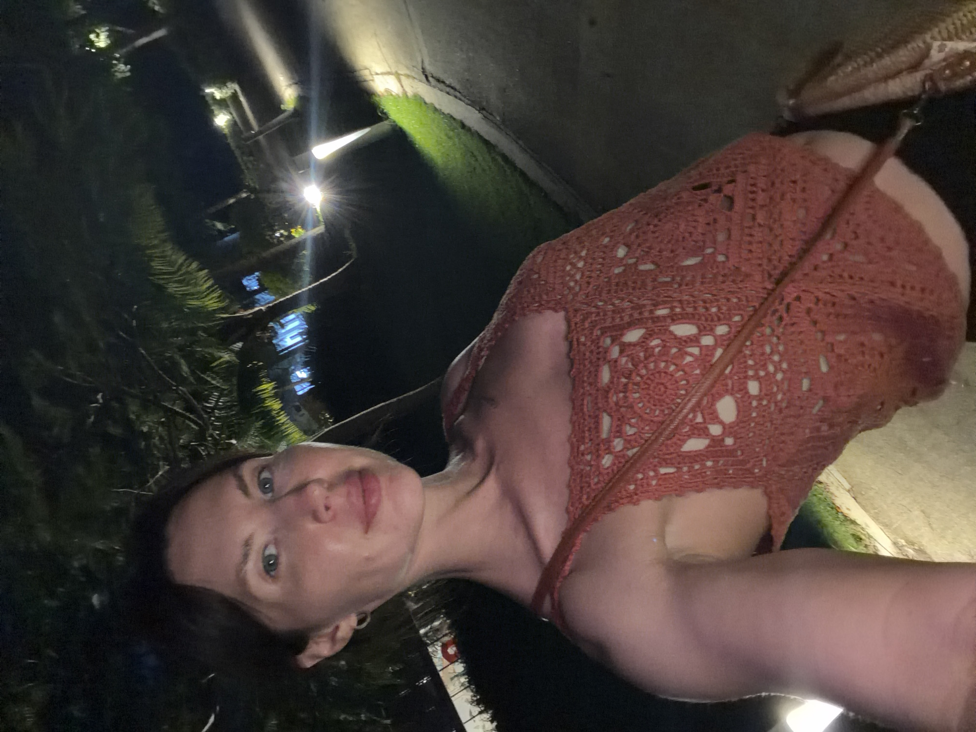 A woman is standing outdoors at night, illuminated by artificial lights. She is wearing a coral-colored crocheted top and a cross-body bag. In the background, there are palm trees, a paved walkway, and bright lights, suggesting a resort or outdoor dining area. The location is identified as Dong Ninh Hoa Ward, Vietnam.