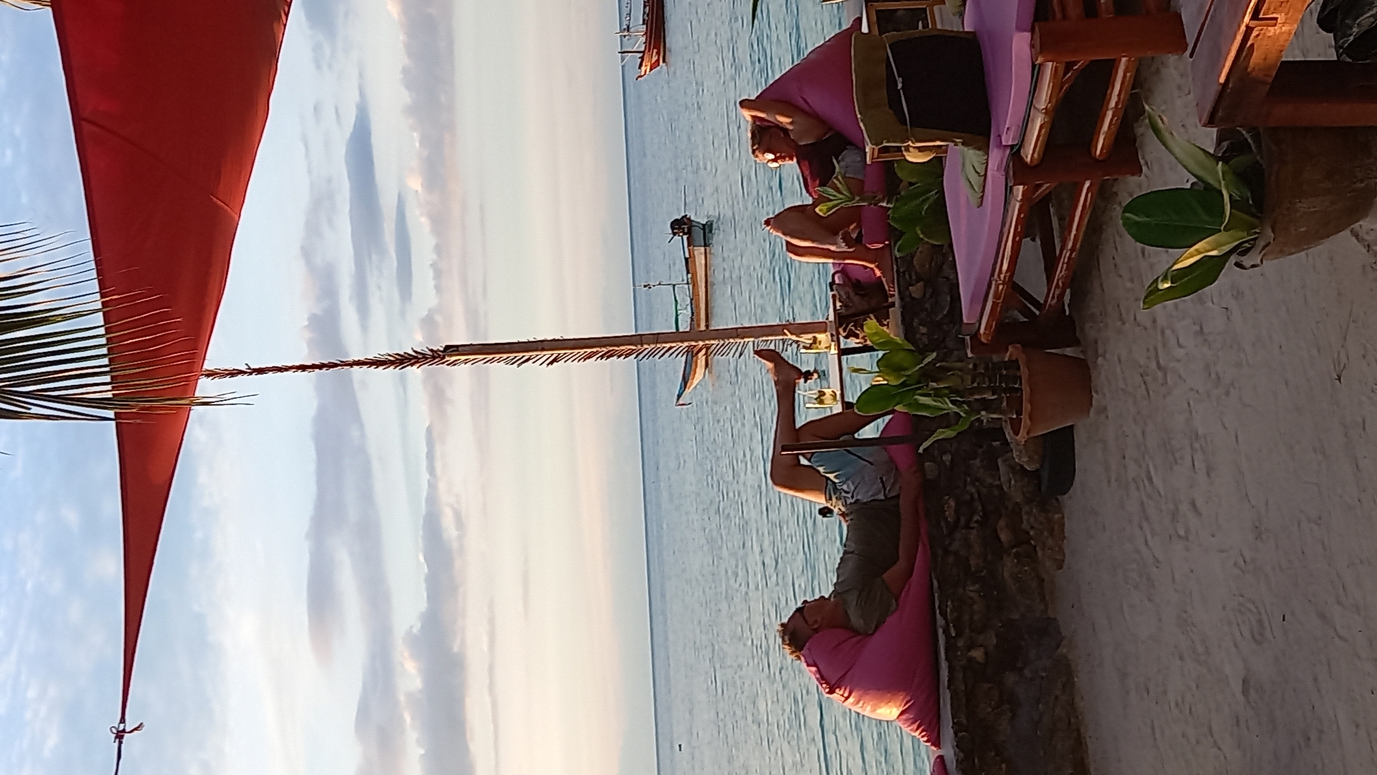 A person is relaxing on a pink beanbag chair on a beach. The beanbag chair is placed on a sandy surface, and behind it, a wooden table and chairs are visible. The table has some greenery on it. The person is wearing shorts and a t-shirt and appears to be enjoying the view of the ocean. In the background, the calm sea stretches out to the horizon, under a sky with scattered clouds. The sun is low in the sky, casting a warm glow over the scene, suggesting it is either sunrise or sunset. A red canopy is partially visible in the foreground, providing shade. The overall atmosphere is one of relaxation and tranquility. The location is specified as Koh Phangan, Thailand.