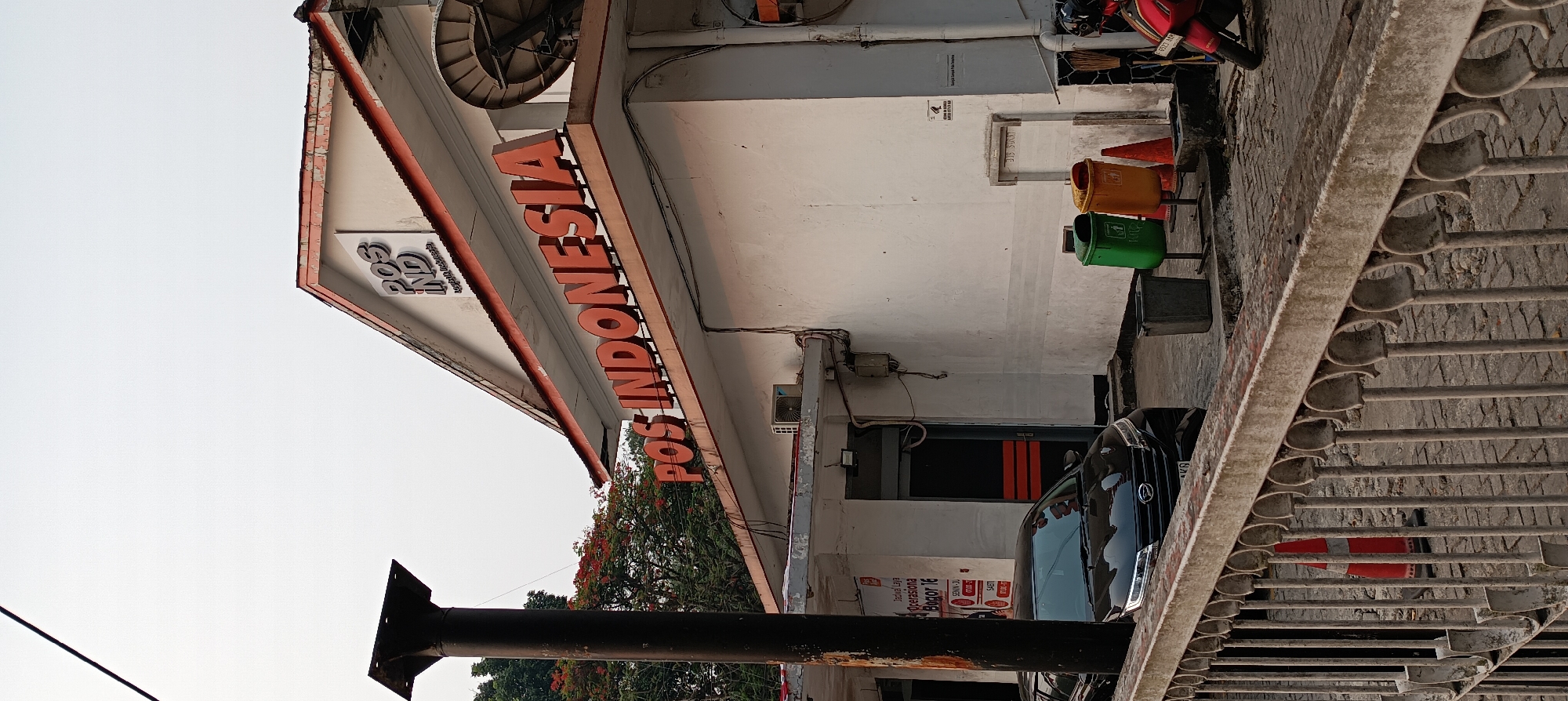 The image shows the front of a Pos Indonesia building, a postal service in Bogor, Indonesia. The building has a white facade with orange trim and signage. Prominently displayed is the "POS INDONESIA" logo in large orange letters. Above this, smaller lettering reads "POS INDONESIA". To the left of the building, a black car is parked. Near the entrance, there are two trash bins, one green and one orange, and an orange traffic cone. In the background, trees with red foliage are visible, suggesting it might be the autumn season. A satellite dish is mounted on the side of the building. A weathered black pole stands tall to the left of the frame, obscuring part of the building. The ground is paved with cobblestones. A metal railing runs along the bottom of the image. The sky is overcast and pale.