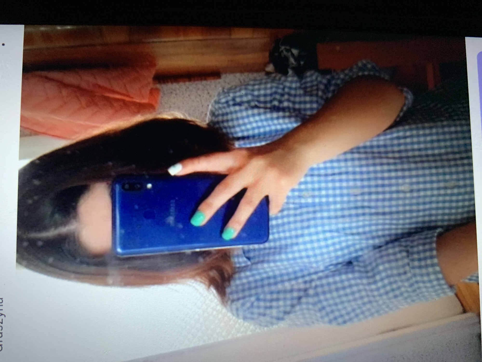The image shows a person, likely a woman, taking a selfie in a mirror. She is wearing a blue and white checkered shirt. Her fingernails are painted a bright turquoise color. She is holding a blue smartphone in her right hand, which is obscuring part of her face. Her left hand is raised, showing two fingers in a peace sign. The background is somewhat cluttered, with a folded orange fabric item hanging on the right and what appears to be a wooden surface. The lighting suggests an indoor setting, possibly a home. There is no indication of the city of Lymanka, Ukraine, or any other specific location.