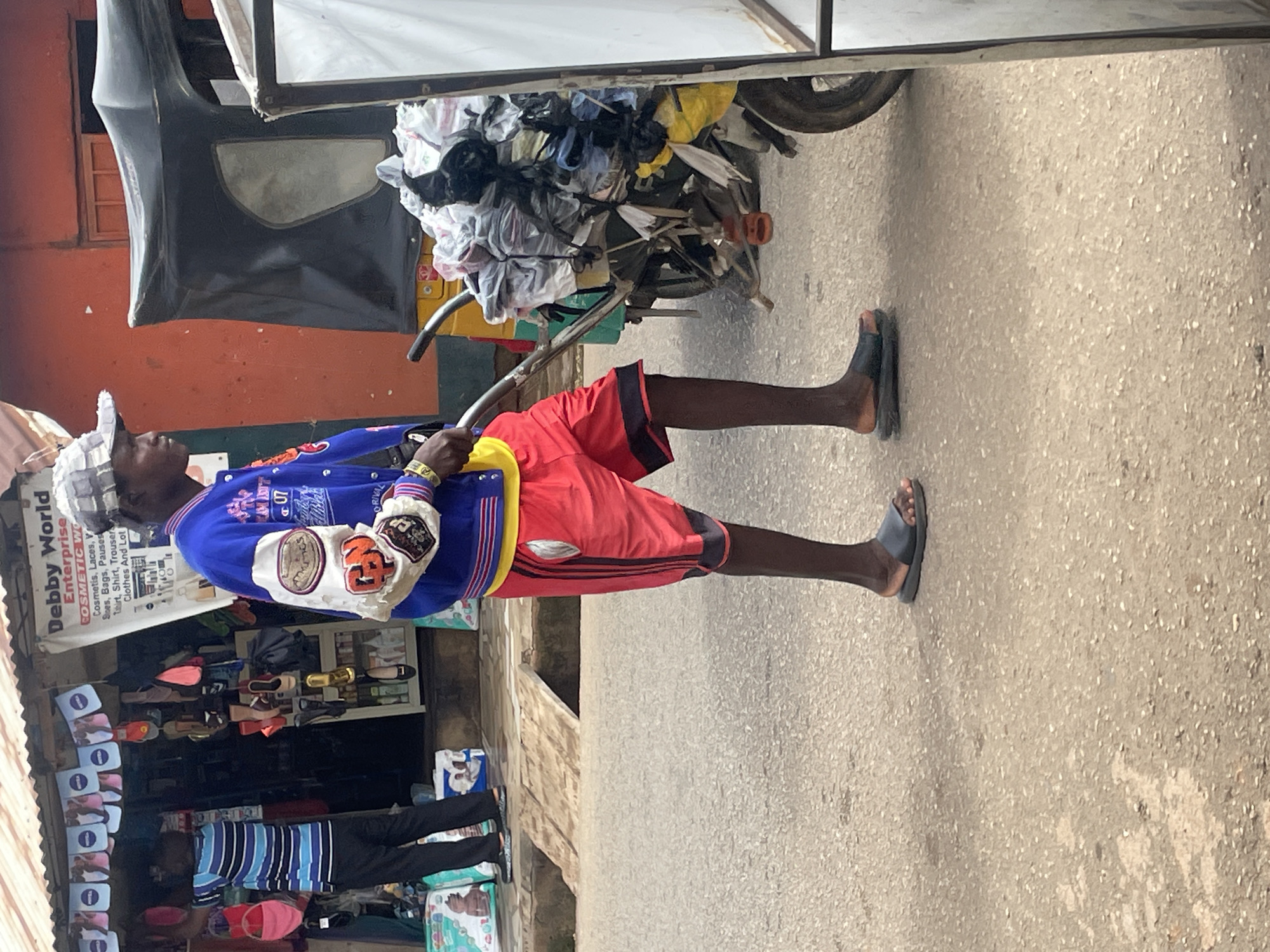 A full-shot, eye-level, outdoor vertical image shows a man in a blue jacket and red shorts pushing a cart filled with bags and miscellaneous items. The man, who is the central focus, is wearing a grey and white checkered cap, a blue letterman-style jacket with patches, a yellow shirt underneath, and red shorts with black trim. He has on black sandals. He is walking on a paved surface with a slight incline. Behind him, to the left, is a shop with a red-orange exterior and a white awning. The shop displays various items on shelves and hanging from the ceiling. A sign above the shop entrance reads "Debby World Enterprise" and lists cosmetic products, shoes, bags, and clothing. Another person, wearing a blue and white striped shirt and black pants, is visible inside the shop. To the right of the man and his cart is a portion of a black, three-wheeled vehicle, possibly a tuk-tuk. The background shows a red-orange wall and a street. The environment appears to be an urban setting in Gura Top, Nigeria, with the shop and cart suggesting a commercial street. The lighting is bright, indicating it is daytime.