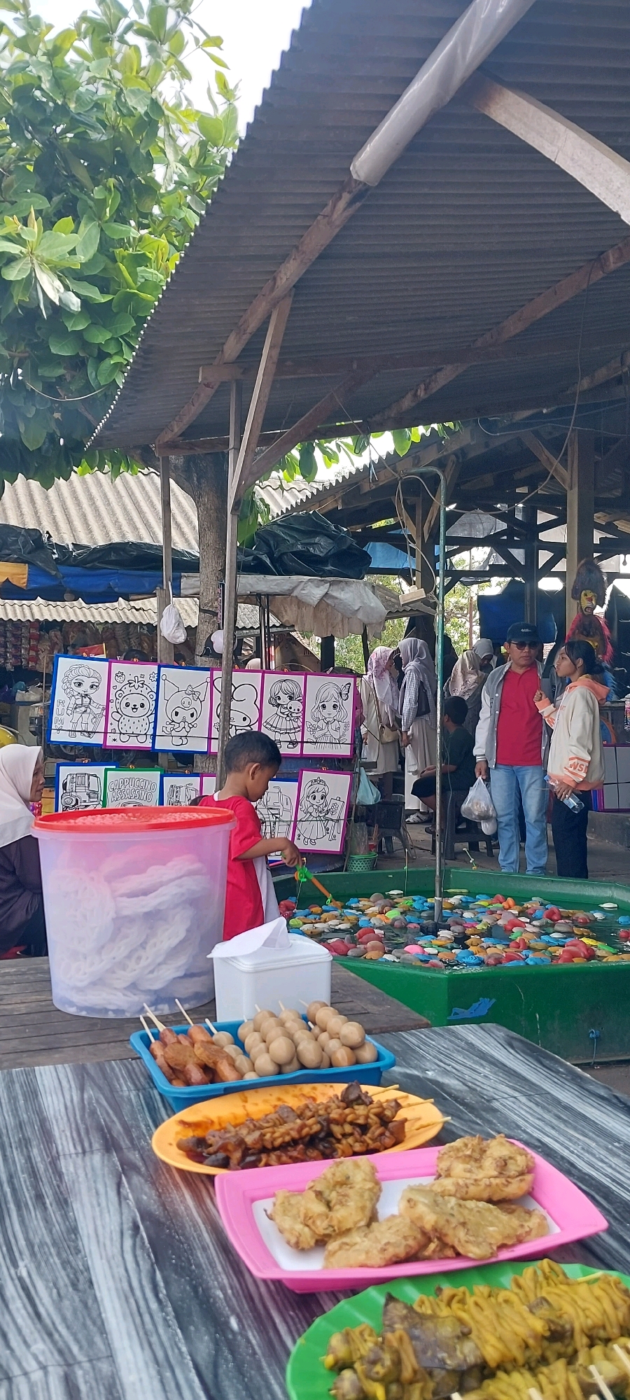 In Pecangaan, Indonesia, this image captures a bustling outdoor market scene. A child wearing a red shirt is actively engaged in a fishing game, using a rod to try and catch colorful toy fish floating in a green pool. Behind the child, a large transparent plastic container is filled with white, airy snacks, likely crackers. In the foreground, a wooden table displays an assortment of Indonesian street food: skewers of meat and meatballs in blue and yellow trays, a pink tray with fried food items, and a green tray filled with yellow seasoned skewers. The market is partially covered by a rustic, corrugated metal roof supported by wooden beams, with leafy trees extending from the top left. Several other individuals are visible in the background, some browsing stalls or walking through the market. The atmosphere suggests a lively community gathering or local festival.