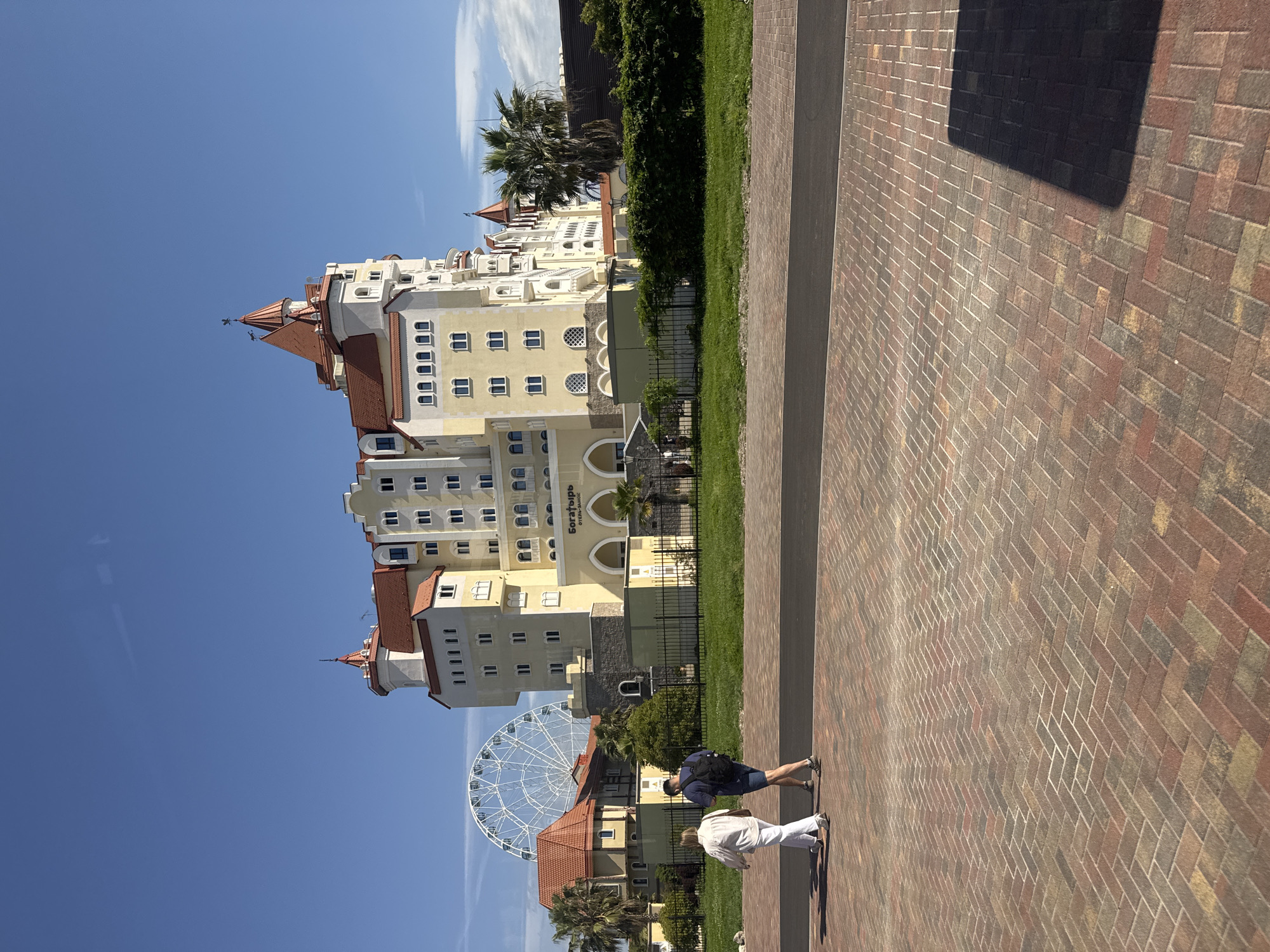 The image depicts a building and its surrounding area in Sirius, Russia.
**Setting and Environment:**
A large, multi-story building, possibly a hotel or resort, dominates the scene. It features light-colored walls, red roofs, and architectural elements reminiscent of a fairytale castle. In the background and to the left of the building is a ferris wheel, which suggests the presence of an amusement park or entertainment area. The building is adjacent to a walkway paved with red bricks. A strip of grass and some foliage are visible between the building and the walkway.
**People:**
Two people are walking on the brick walkway. One is wearing a white outfit, and the other is wearing a dark blue shirt with dark shorts.
**Objects:**
The image contains the building, the ferris wheel, a brick walkway, grassy areas, and foliage. There are also fences around the base of the building.
**Actions and Interactions:**
The two people are walking along the walkway. The building is standing still.
**Overall Impression:**
The image presents a vibrant, tourist-friendly environment in Sirius, Russia, with a unique blend of architectural design and recreational attractions. The photograph suggests a pleasant, sunny day.