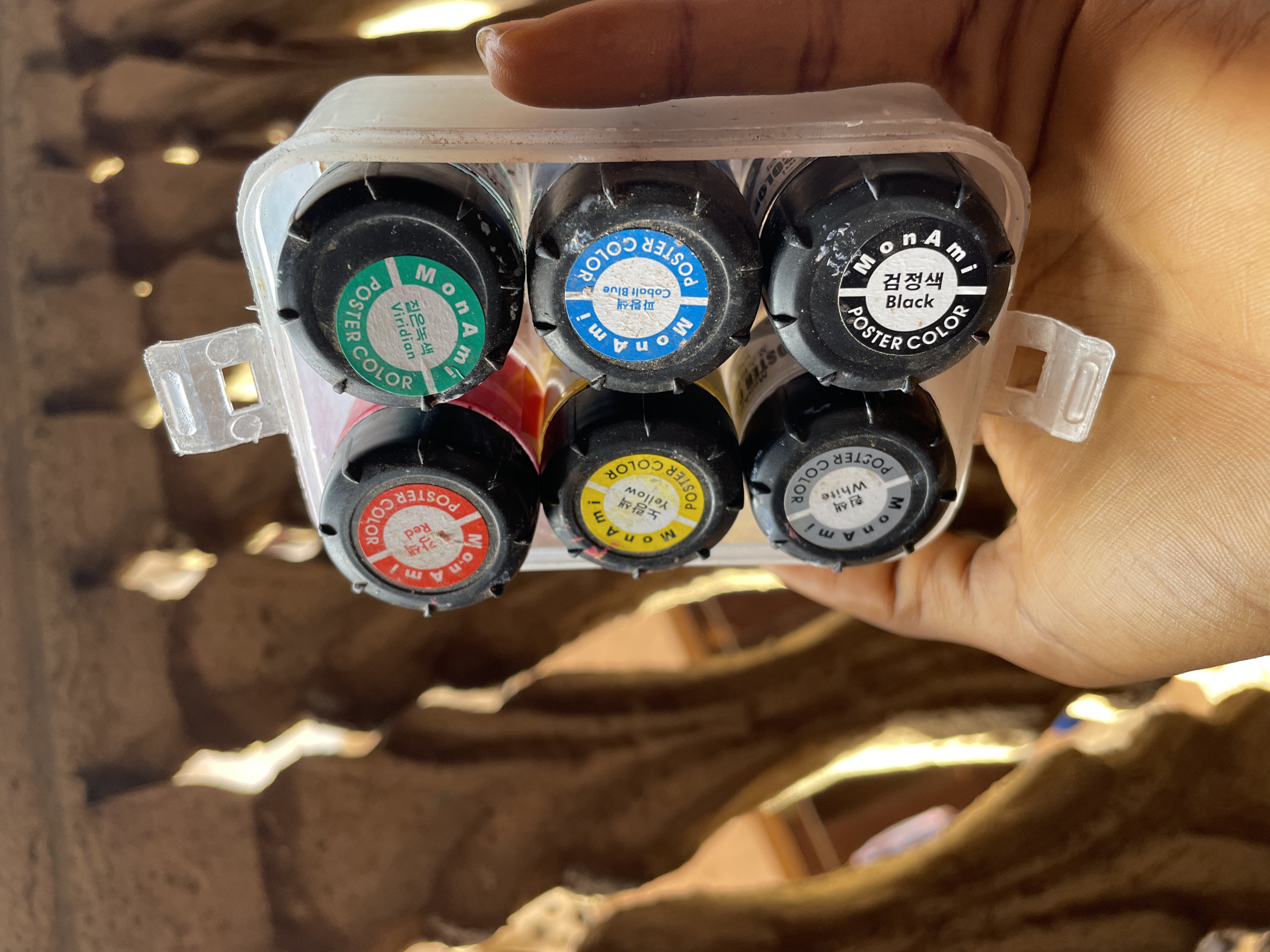 A person is holding a clear plastic container holding six bottles of MonAmi poster paint. The colors, labeled in English and Korean, include: Viridian, Cobalt Blue, Black, Red, Yellow, and White. The background is blurry and indistinct but appears to be a neutral brown color. The image does not contain sufficient information to identify Nkpor, Nigeria.