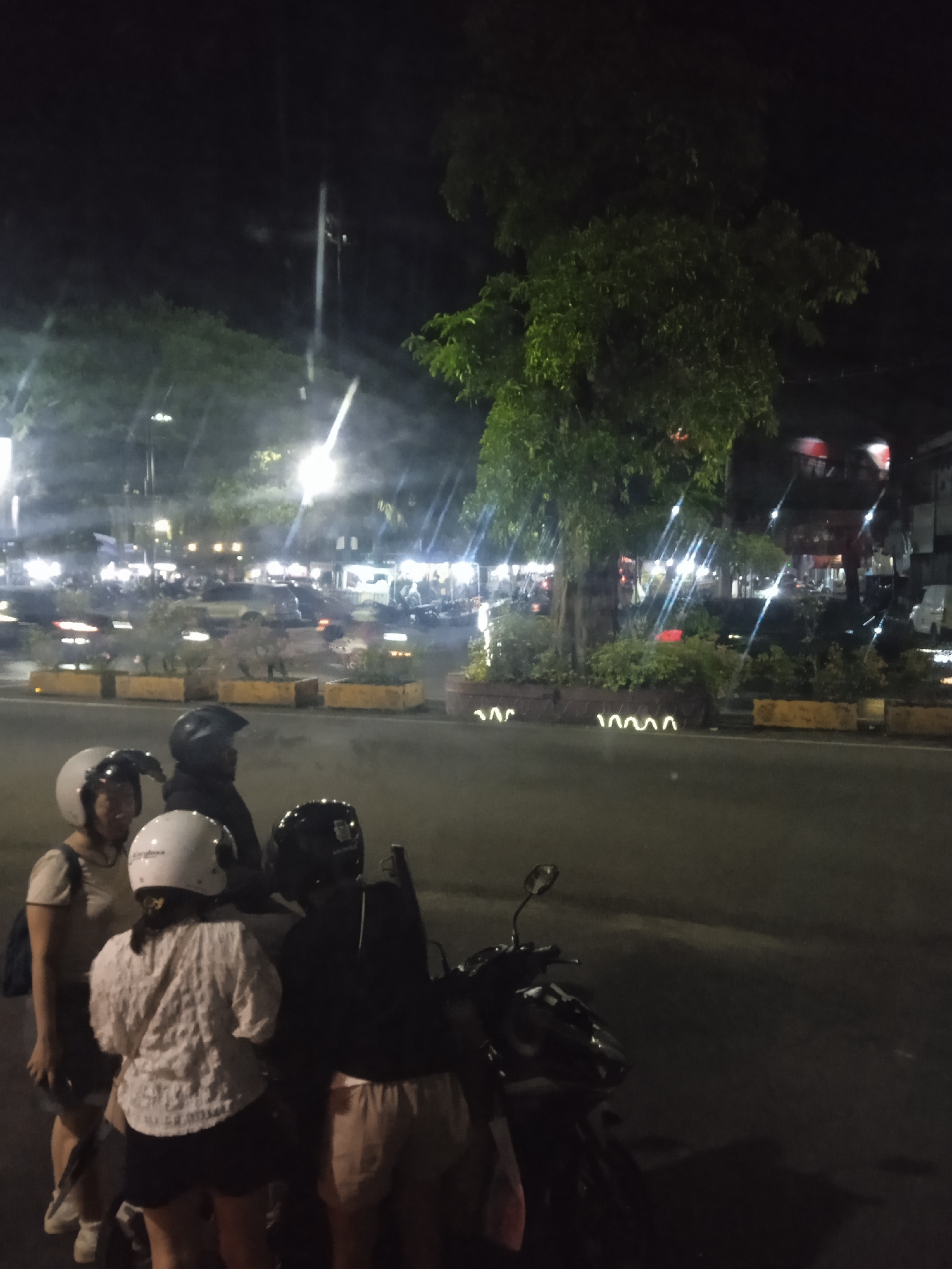 The image depicts a night scene in Madiun, Indonesia. Four people wearing helmets are gathered around a parked motorcycle on a street with a median. The median has plants and lighting fixtures. In the background are streetlights and cars. Buildings line the background.