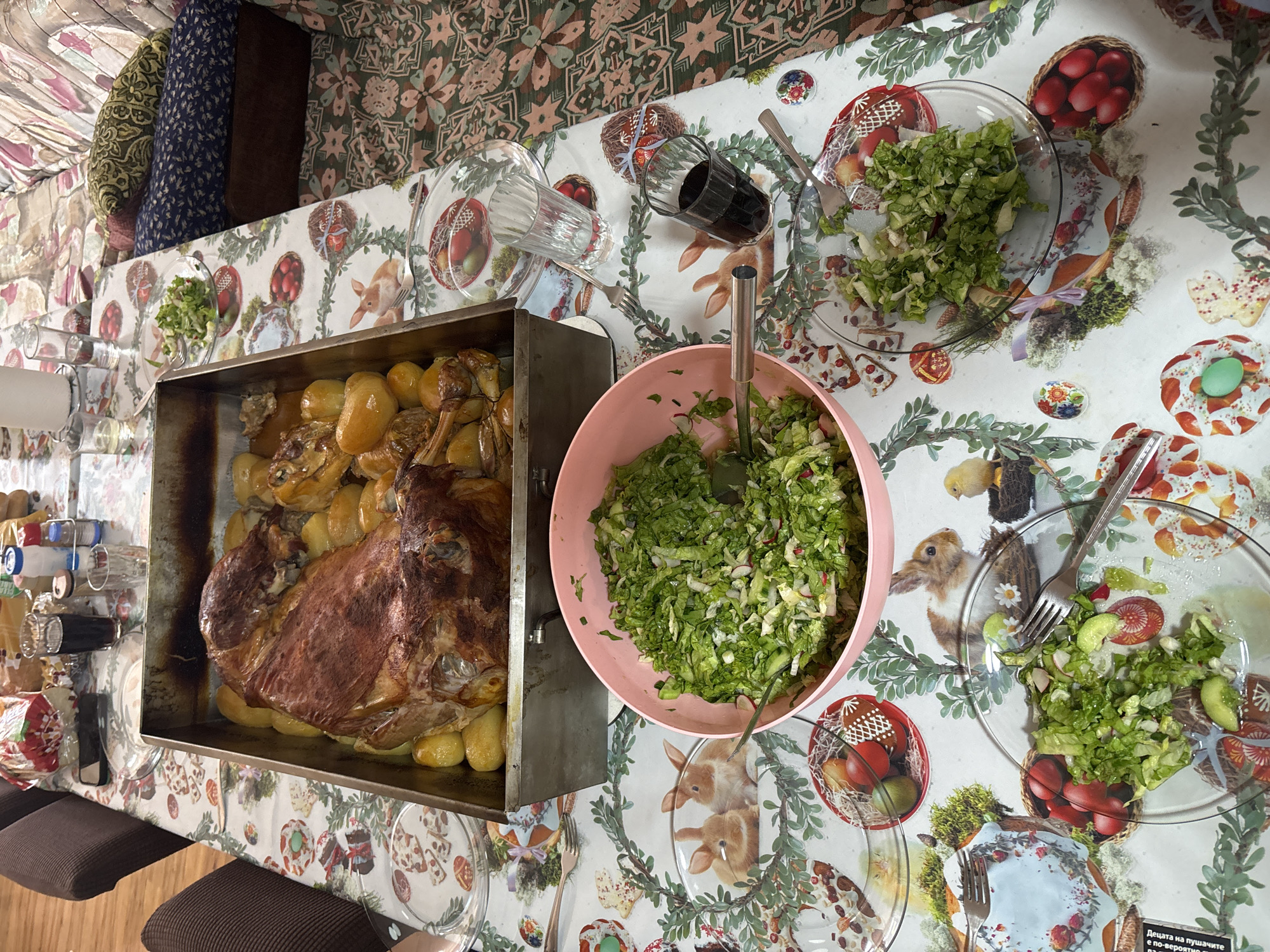 The image shows a table setting, likely indoors, featuring a roasted leg of lamb with potatoes in a large metal pan, a large pink bowl filled with salad, and individual salad plates. A glass of dark liquid (possibly wine or juice) is also visible, along with other glasses and utensils. The tablecloth has a festive, possibly Easter-themed, design with bunnies and eggs. The image was taken indoors, and the location, Novo Zhelezare, Bulgaria, suggests this may be a family meal or celebration in a home setting.