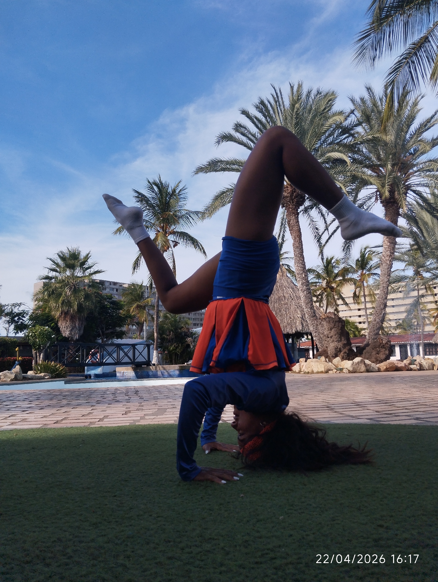 A person is captured performing a headstand or handstand on green artificial grass in an outdoor, sunny environment. The individual is wearing a blue long-sleeved top, an orange and blue pleated skirt over blue shorts, and white socks. Their dark hair cascades onto the grass, and their legs are bent and raised into the air, indicating an acrobatic pose.
The background reveals a resort-like setting with a paved area, a blue swimming pool or water feature, and a decorative bridge with an X-patterned railing. Numerous tall palm trees dominate the midground and background, interspersed with multi-story buildings that suggest a hotel or residential complex. A small hut with a thatched roof is also visible. The sky above is clear blue with a few wispy white clouds, indicating pleasant weather. The sunlight suggests it is daytime, confirmed by the timestamp "22/04/2026 16:17" displayed in the bottom right corner of the image.