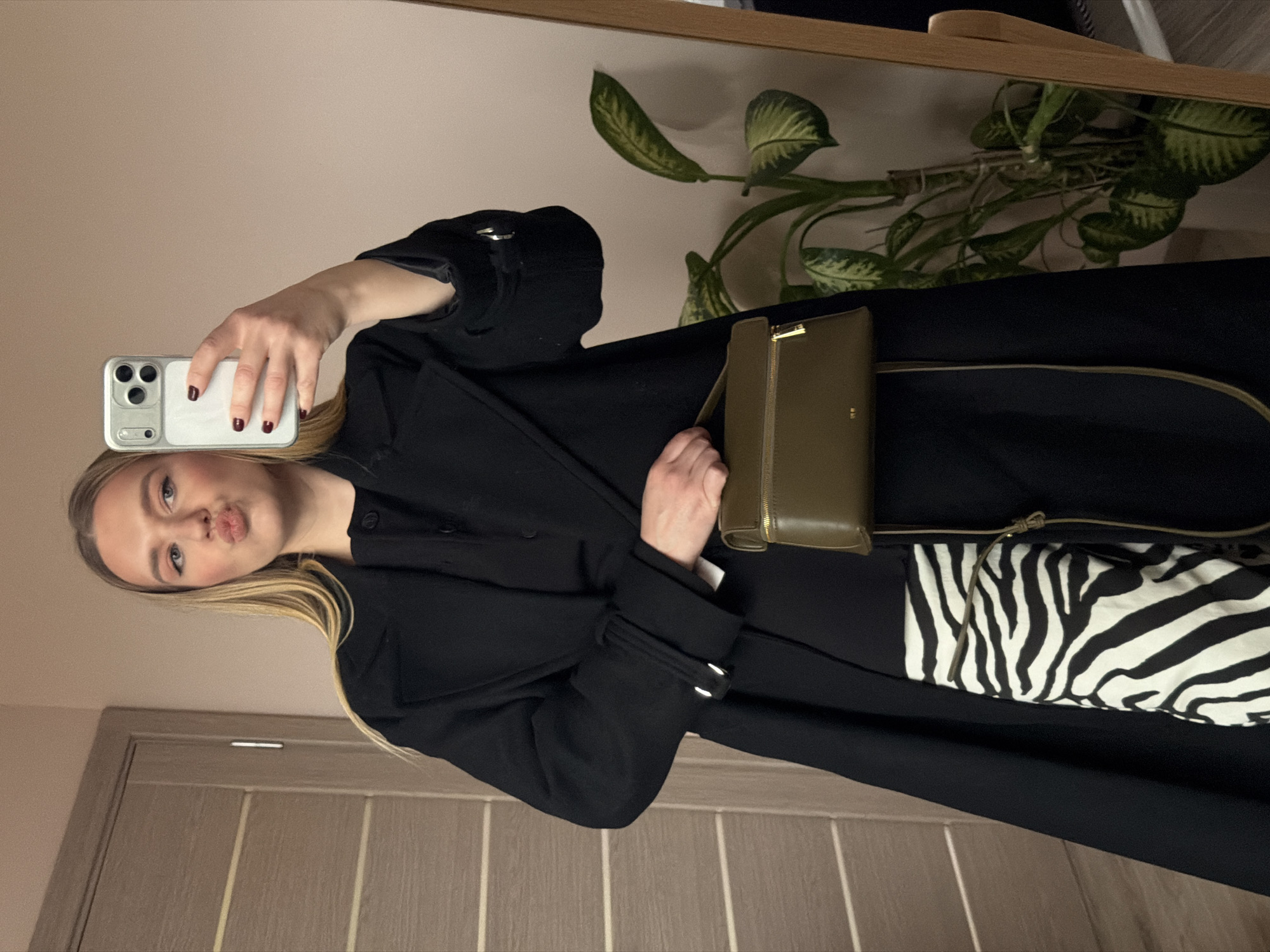 A woman is taking a selfie in a mirror. She is wearing a black coat and carrying a small, olive green purse. Part of a zebra-print garment is visible. A houseplant sits on a surface behind her. A beige door is partially visible to the left. The background suggests an indoor setting. Dreiliņi, Latvia is not discernible from the provided image.
