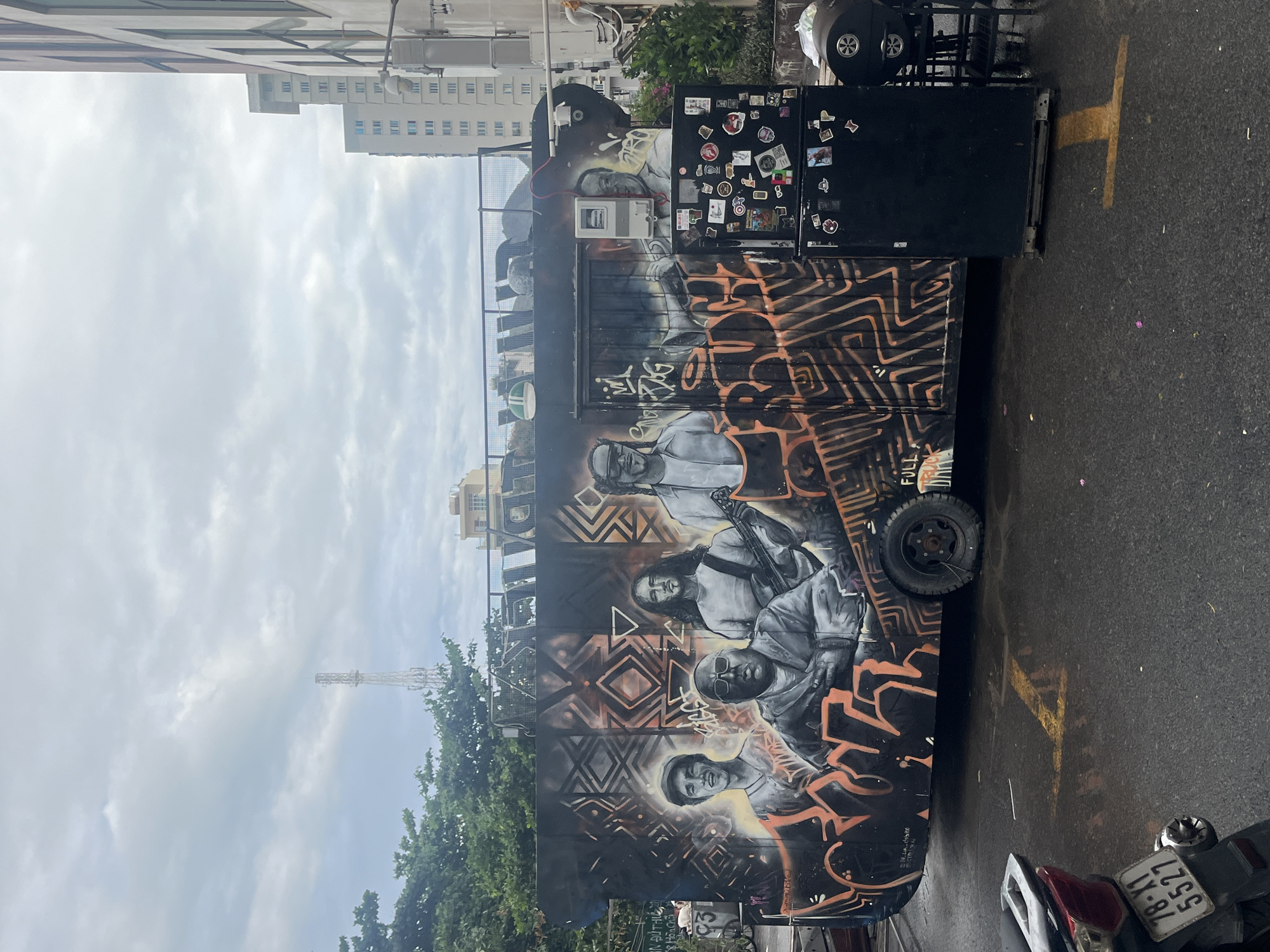 A street in Bac Nha Trang Ward, Vietnam, displays a mobile food stall adorned with graffiti art. The artwork depicts four individuals in a black and white palette, integrated into a backdrop of abstract patterns in black and orange. The stall itself has a small black refrigerator covered with stickers. A motorcycle is parked nearby. The sky is overcast, and buildings are visible in the background.