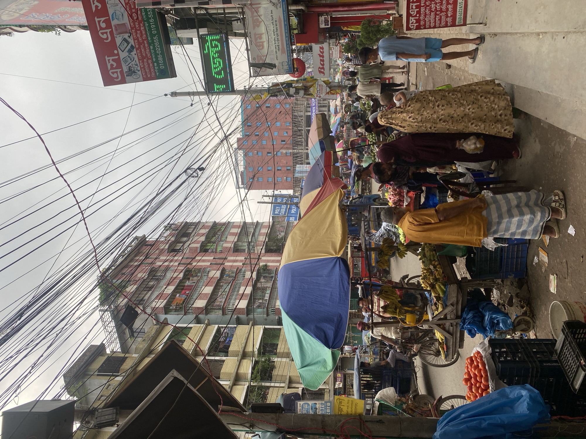 The image depicts a street scene in Dhaka, Bangladesh, characterized by a bustling marketplace and dense overhead electrical wires. In the foreground, a fruit stall displays a large pile of tomatoes in a crate, alongside bunches of bananas. Several people are visible, including a man in an orange shirt and striped lungi, a woman in a maroon hijab, and another woman in a patterned hijab carrying a baby. In the background, a multi-story building with red and white striped facade stands prominently, interspersed with other buildings and street signs. Numerous motorcycles and carts are also present, suggesting a busy commercial area. The sky is overcast, and the overall impression is one of a vibrant, somewhat chaotic urban environment.