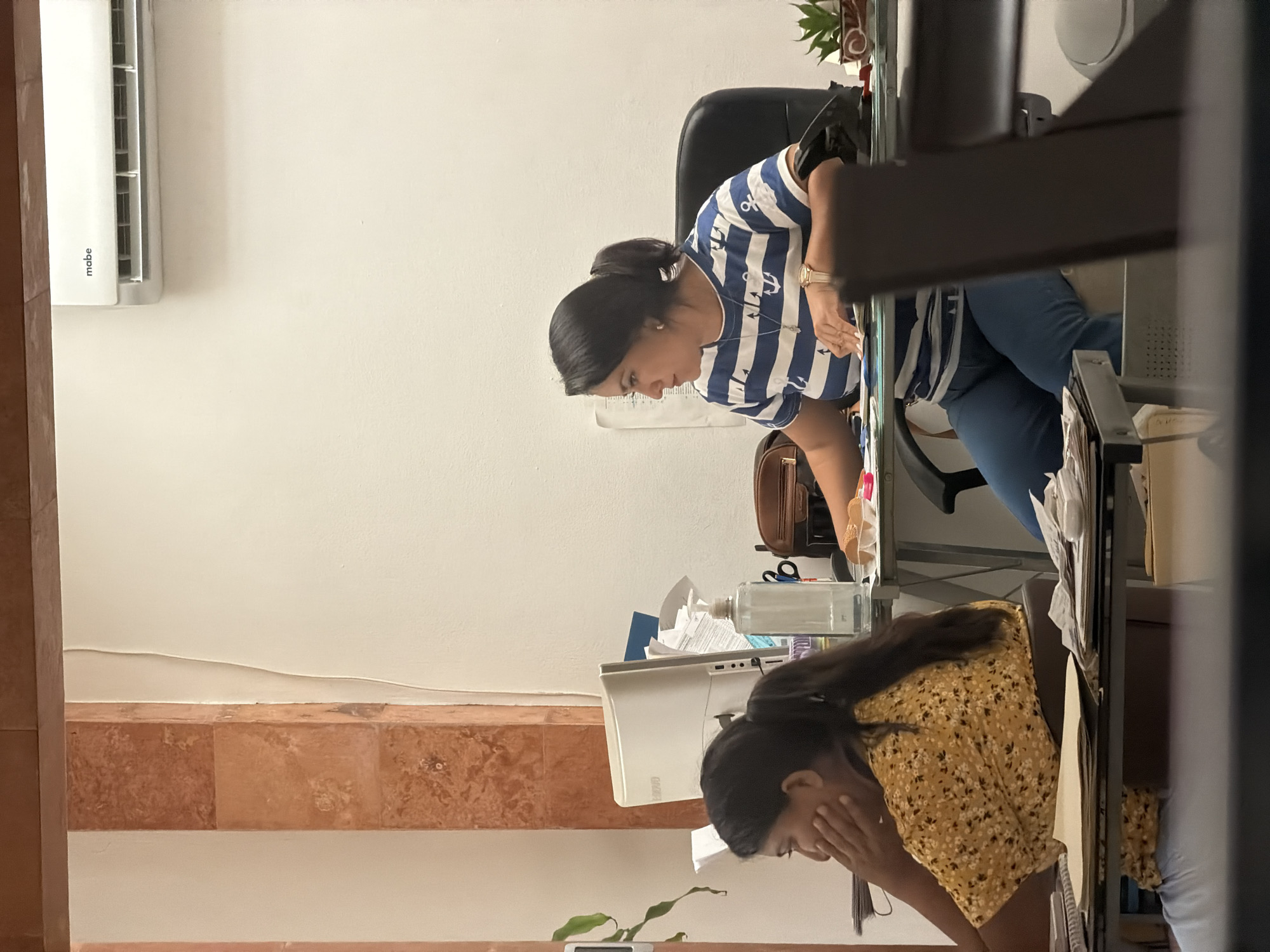 The image depicts an indoor scene, likely an office environment in Cancún, Mexico. Two women are seated at desks. The woman on the right is wearing a blue and white striped shirt with anchor patterns and blue pants. She has dark hair pulled back in a ponytail and is looking down at papers on her desk, with a watch on her left wrist. To her left is a computer monitor, a hand sanitizer dispenser, and a brown leather bag. A small potted plant sits to the right of her desk.
The woman on the left is wearing a yellow floral print shirt and light blue jeans. She has dark hair tied in a ponytail and is resting her chin on her hand, looking to the left. Papers are spread across her desk in front of her.
The environment appears functional, with office equipment and documents visible. The wall is white, and there's a beige and reddish-brown decorative element on the left side of the frame, possibly a wall accent. An air conditioning unit is mounted high on the wall. The lighting suggests it is daytime. The overall impression is of a working office.