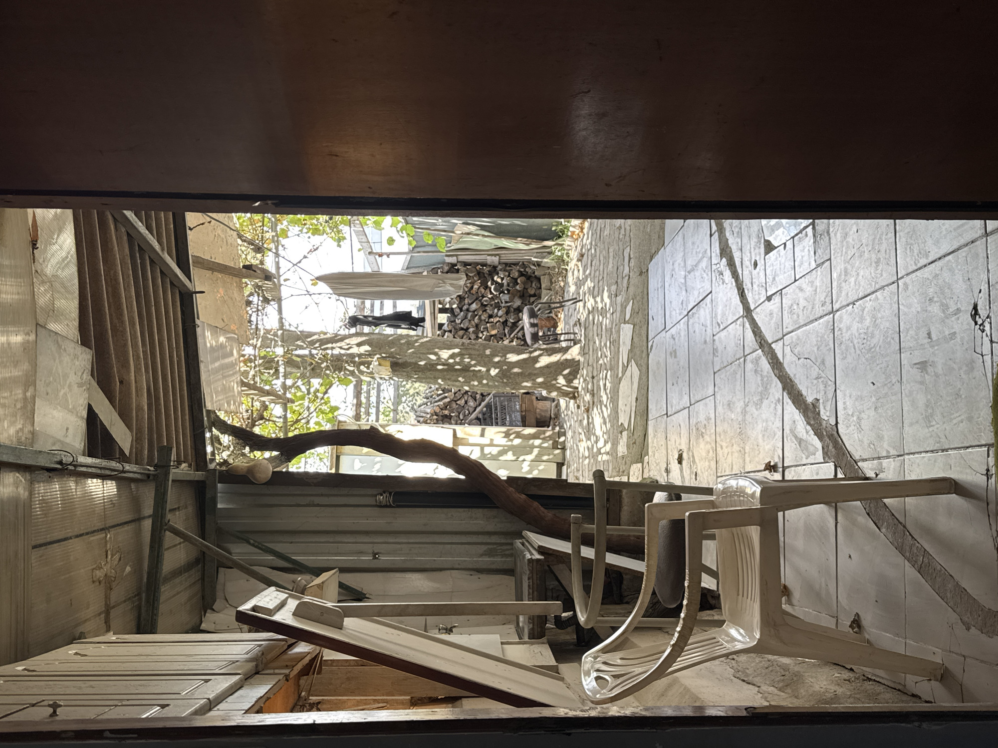 The image depicts a rear outdoor area viewed from an open doorway. In the foreground, two white plastic chairs with grey cushions are placed on a tiled patio. The patio appears weathered, with some cracks and debris scattered on it. To the left, a corrugated metal wall is visible, with some wooden elements and a tilted mirror leaning against it.
Beyond the patio, the scene opens up to a yard with a large tree whose trunk is gnarled and leans significantly. The ground in this area is paved with irregularly shaped stones or bricks. Behind the tree, a pile of cut firewood is stacked. A small, dark stool is positioned near the tree. Clothes are hanging on a line in the background, along with a light-colored fabric sheet. The overall environment suggests a somewhat rustic and utilitarian outdoor space, possibly a backyard or a service area. The lighting indicates it is daytime, with sunlight casting distinct shadows from the tree. The location is identified as Gebze, Turkey.