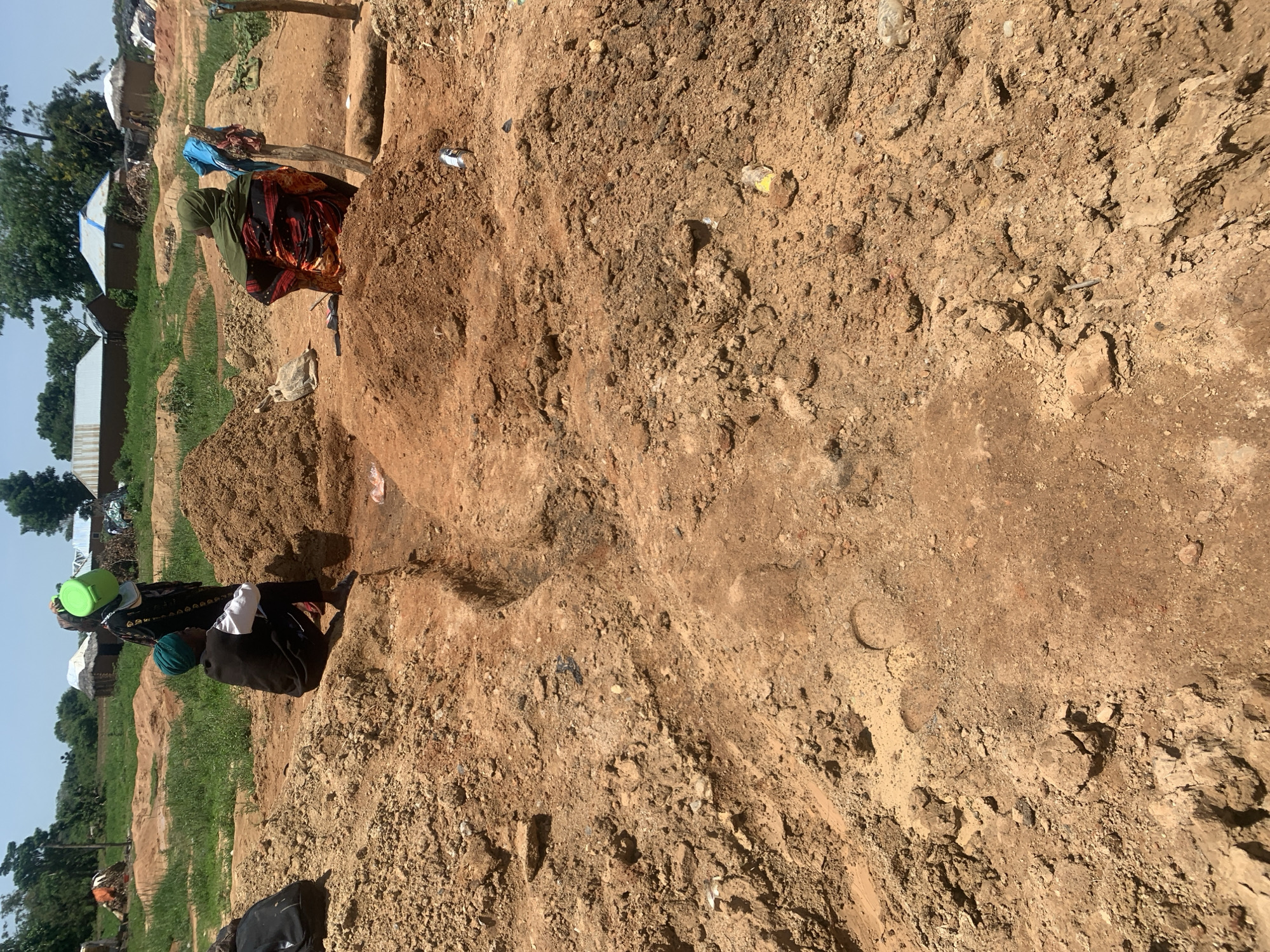 In Buga da Gindi, Nigeria, a scene unfolds amidst dry, dusty terrain. Three individuals are present, engaged in activities that suggest manual labor or daily life. One person, dressed in a black vest and green headscarf, is seated on a mound of reddish-brown soil, facing left. Another person, wearing a black garment with a green head covering, stands near a bright green bucket. The third individual, adorned in a colorful patterned dress and green headscarf, is also seated, facing away from the camera.
The environment is characterized by uneven, disturbed ground, with piles of excavated earth. In the background, simple structures with thatched or corrugated metal roofs are visible, indicative of a rural or semi-rural settlement. The overall impression is one of an outdoor work area or a communal gathering space in a dry climate. Scattered debris, including plastic and metal containers, are also present, suggesting human activity.