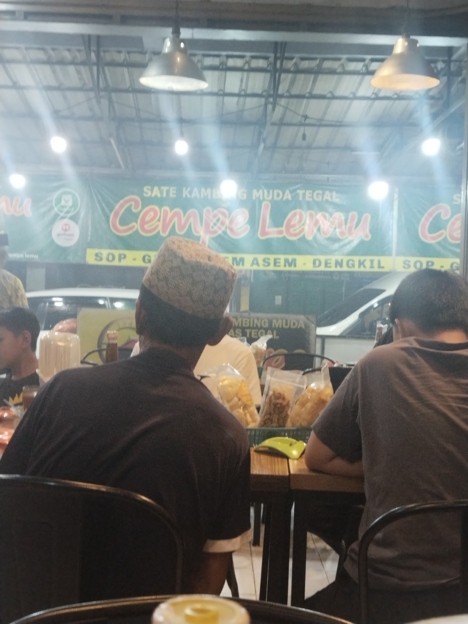 The image depicts an indoor scene, likely a food establishment, possibly in Adiwerna, Indonesia, given the context provided. The setting appears to be casual, with several people seated at tables. In the foreground, two men are visible from behind, seated at a wooden table. One man on the left wears a dark brown long-sleeved shirt and a patterned cap. The man on the right wears a dark grey t-shirt. Bags of snacks or food items are on the table between them. In the background, a large green banner hangs on the wall with text that reads "SATE KAMBING MUDA TEGAL CEMPE LEMU". Various light fixtures are visible on the ceiling. A white vehicle can be seen through a window or opening in the background. The overall atmosphere suggests a busy, local eatery.