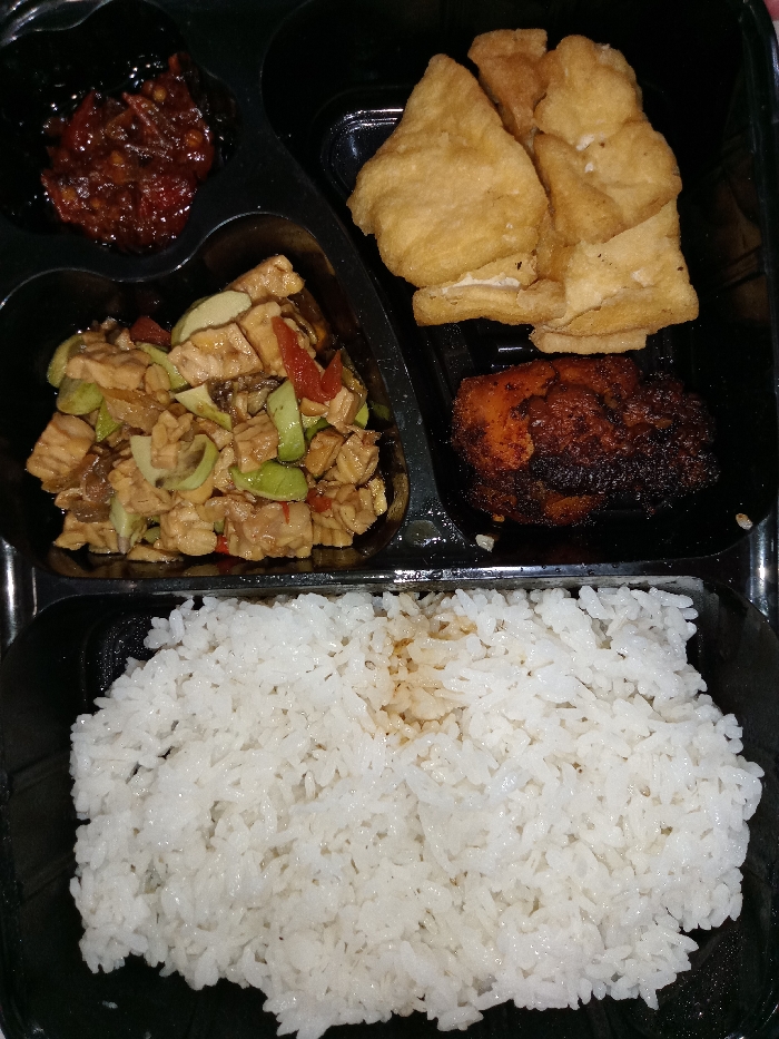 This media file displays an aerial view of a ready-to-eat meal presented in a black, partitioned takeaway container, indicative of Indonesian cuisine. The meal is divided into several compartments.
In the top-left compartment, there is a small serving of dark red sambal or chili paste, featuring visible chili flakes and possibly other spices, suggesting a spicy condiment.
The middle-left compartment holds a stir-fried dish, predominantly consisting of cubed tempeh and sliced green petai (stink beans). Red chili slices are also interspersed throughout the dish, which is coated in a dark, glossy sauce.
In the top-right section, several pieces of fried tofu are neatly stacked. The tofu has a light golden-brown exterior, indicating it has been deep-fried.
Below the fried tofu, in the bottom-right small compartment, a piece of chicken or meat, possibly grilled or fried, is visible. It has a rich, dark brown color with some charred edges, suggesting a flavorful and well-cooked preparation.
The largest compartment at the bottom of the container is filled with a generous portion of plain white rice. Some light brown liquid or sauce stains are visible on the surface of the rice, possibly from one of the other dishes.
The scene depicts a typical Indonesian meal packed for convenience, likely for lunch or dinner. The food appears freshly prepared, and the black container provides a stark contrast to the colorful dishes. No text is visible in the image. Given the location context of Situbondo, Indonesia, these dishes are characteristic of local fare.