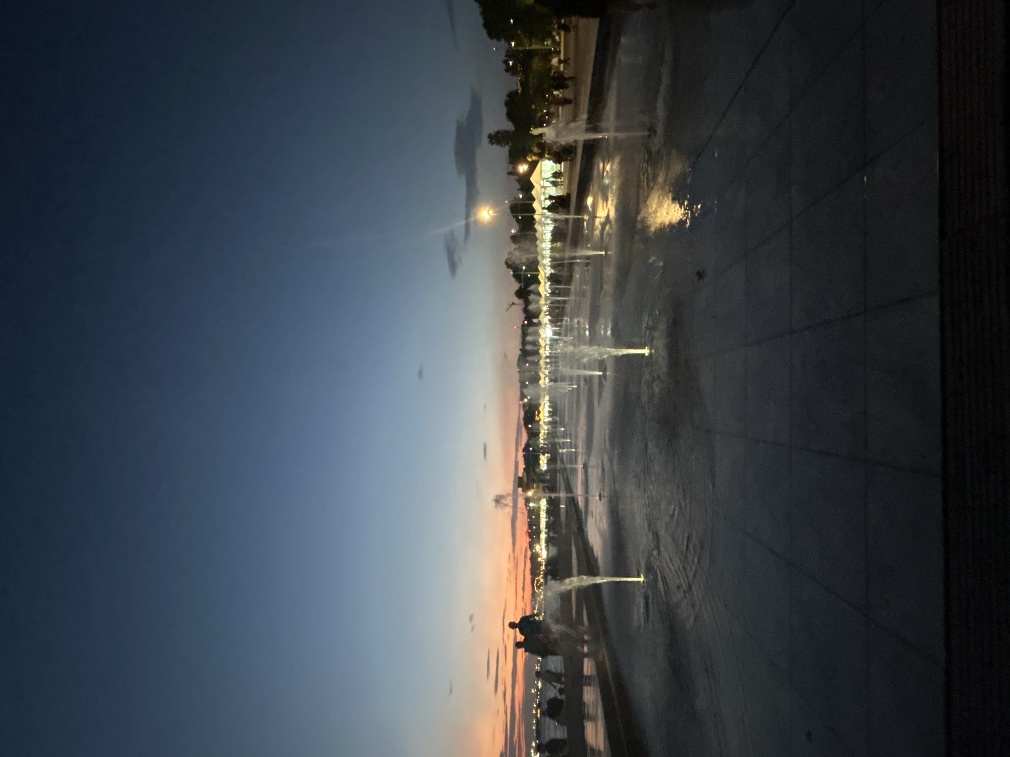 This is a nighttime photograph taken at the Thessaloniki Municipal Unit, Greece, likely at the waterfront promenade known as Nea Paralia. The image captures a wide-angle view of an outdoor public space with numerous illuminated fountains. The sky is a deep blue, transitioning to warmer hues of orange and pink near the horizon, indicating dusk or twilight.
In the foreground, a paved area with a grid pattern is wet from the fountains, with water jets spraying upwards and reflecting the yellow light of the fountains and distant lights. The fountains are arranged in a linear fashion, stretching into the distance.
On the left side of the image, a group of people are visible, silhouetted against the twilight sky. They are standing and sitting near what appears to be a railing or edge of a raised platform, possibly overlooking the water. Some individuals are engaged in conversation, while others are simply enjoying the evening.
In the background, a line of lights and structures suggest a bustling area with possibly shops or cafes along the waterfront. The general ambiance is one of a public gathering place during a pleasant evening. The overall scene evokes a sense of leisure and urban nightlife.