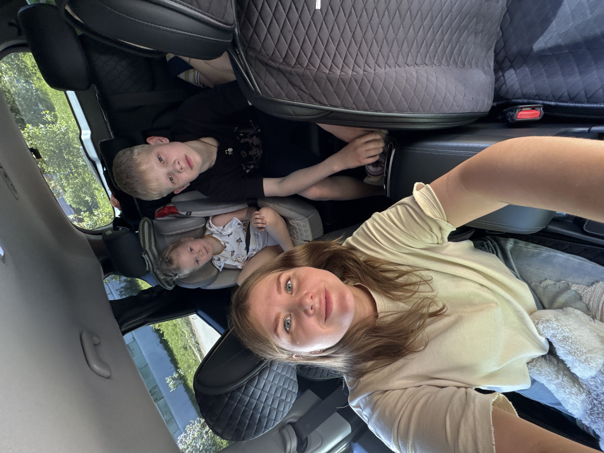Here is a description of the image:
**Scene:** An interior view of a car, likely a passenger vehicle, with three occupants. The photograph is taken from an elevated position, possibly by one of the occupants.
**People:**
* A woman, appearing to be an adult, is in the foreground. She has light brown hair and is wearing a light-colored shirt. She appears to be taking the picture.
* Two children are in the backseat. One is a young boy with short blond hair wearing a black shirt. The other child is younger and seated in a child car seat.
**Objects:**
* Car seats: The younger child is secured in a car seat.
* Car interior: Elements of the car's interior are visible, including upholstered seats, headrests, and parts of the door.
**Environment:** The scene is enclosed within the vehicle, but a blurred view of greenery through the car window suggests an outdoor environment.
There is no information that indicates this photograph was taken in the city of Chaikovsky, Russia.
