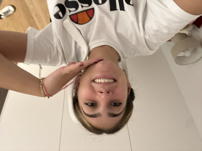 A young woman smiles directly at the camera. She wears a white t-shirt with the "ellesse" logo. Her hair is pulled back, possibly in a towel or headband. Her right hand is raised, with fingertips touching her chin in a casual pose. Bracelets adorn her right wrist. A bedside lamp is visible in the background, along with a glimpse of light-colored flooring. The backdrop consists of white walls.