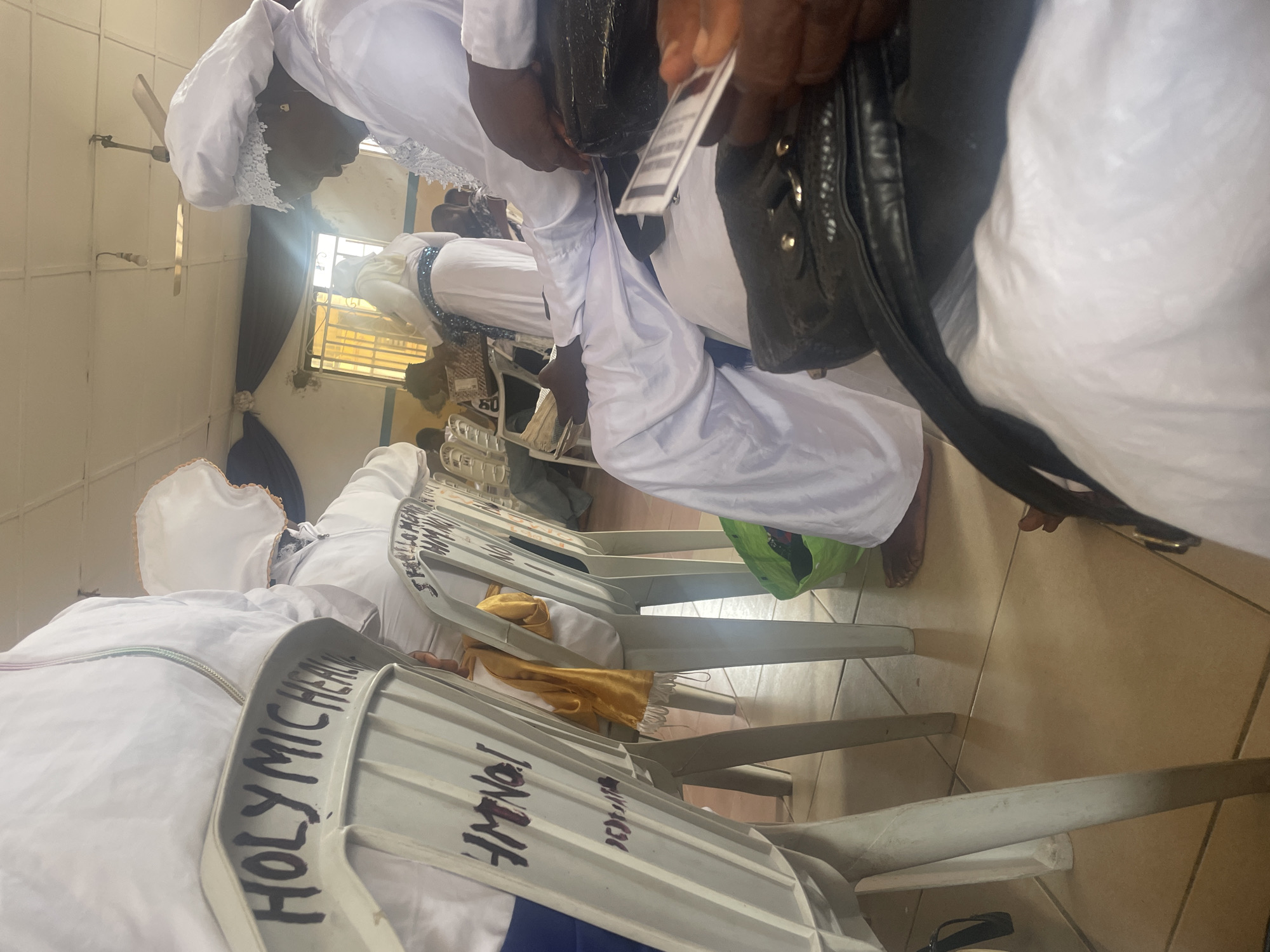 The image shows the interior of a hall, possibly a church or community center, in Lokoja, Nigeria. Several people are seated in white plastic chairs, most of whom are wearing white garments and head coverings, suggesting a religious gathering. The chairs have black lettering on their backs, with visible text such as "HOLY MICHELLE" and "HMNQI." The room has tiled flooring, and a ceiling fan is visible overhead. Natural light streams in from a window on the right side of the image. One person in the foreground is holding a white card with a barcode. A green bag and a yellow cloth are also visible in the scene.