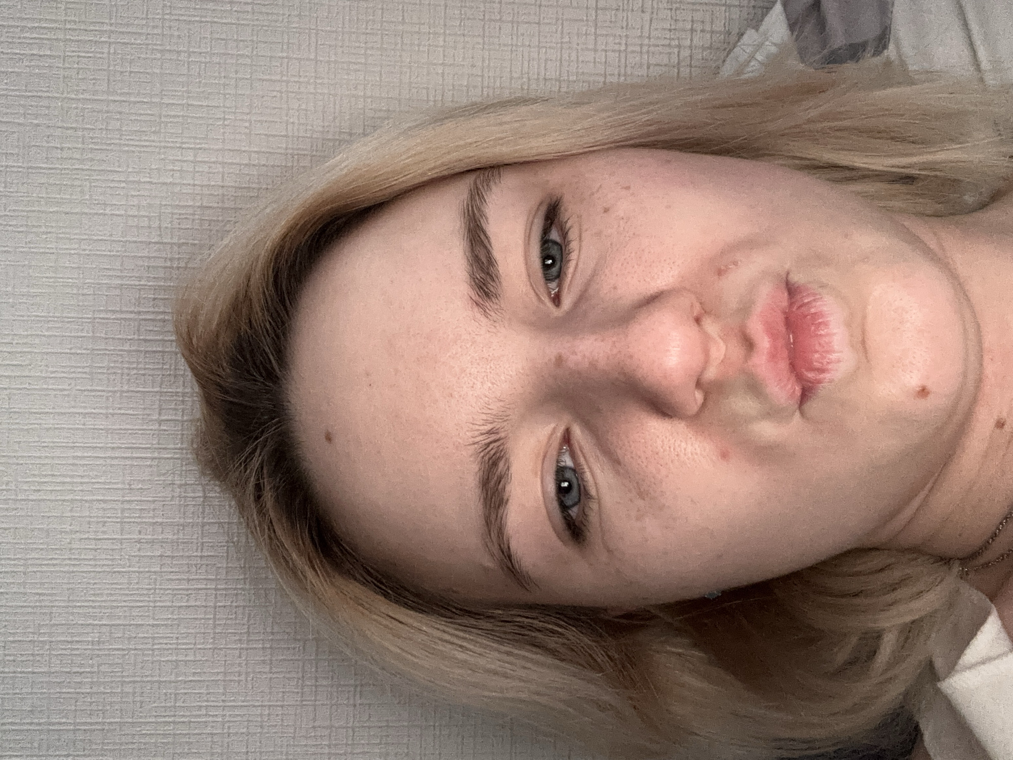 A close-up, tilted headshot depicts a fair-skinned woman with blonde hair and blue eyes. She is making a "duck face" expression, pursing her lips. Her eyebrows are visible and untamed. There are freckles on her face and a mole on her forehead. She appears to be lying down on a light-colored textured surface, possibly a bed. A gray material is also visible in the top right corner of the image, possibly a blanket or pillow. A thin necklace is present near her neck. There is no information in the image that indicates this photo was taken in Vsevolozhsk, Russia.