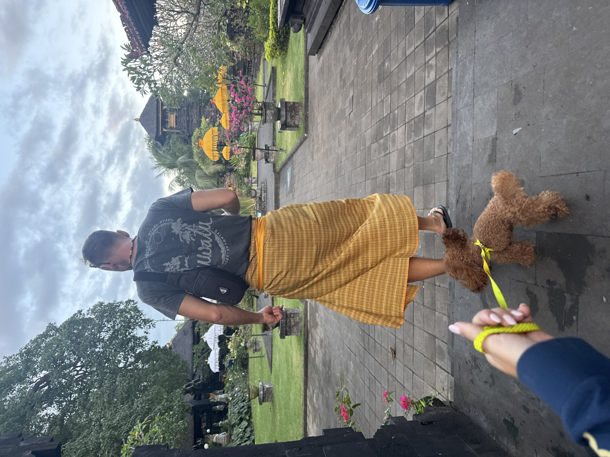 In Dawan, Indonesia, a person from a low-angle perspective holds a leash with a yellow loop, attached to a small brown poodle. The poodle walks on a paved stone path. The leash is held by a person wearing a dark blue jacket with yellow trim. The path leads towards a man wearing a dark t-shirt and a yellow sarong. He is seen from behind, carrying a black bag across his chest. The background shows a lush garden with white umbrellas, pink flowers, and a temple-like structure with a dark roof. The sky is overcast.