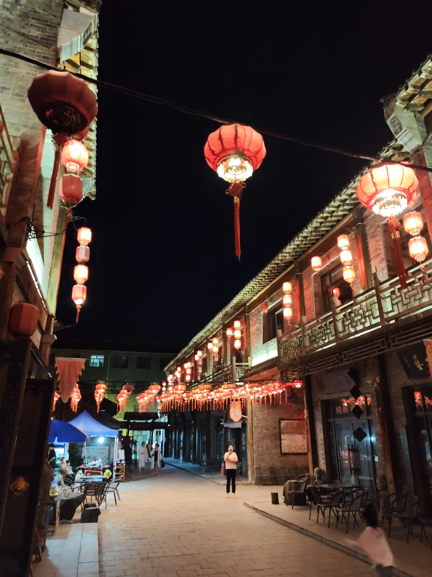 This image depicts a night scene on a street in Lindian, China, adorned with numerous red lanterns. The street is paved with cobblestones and lined with traditional brick buildings that feature ornate wooden balconies and eaves. Red lanterns of various sizes are suspended from wires strung across the street, and also hang from the eaves of the buildings, casting a warm, festive glow. Some lanterns are large and elaborate, while others are smaller and arranged in clusters or strings.

In the foreground, to the left, there is an area with tables and chairs, suggesting an outdoor seating area for a restaurant or cafe. A blue tent is also visible, possibly for a vendor. A few people are present in the image. On the left, near the tent, a person is seated at a table. Further down the street, a woman in a light-colored top and dark pants is standing in the middle of the cobblestone path, holding a phone up as if taking a picture or video. Two figures, dressed in white robes, are walking away from the camera further down the street.

The overall atmosphere is one of a lively, traditional street market or cultural area at night, illuminated by the iconic red lanterns. The dark night sky provides a stark contrast to the bright illumination from the lanterns.