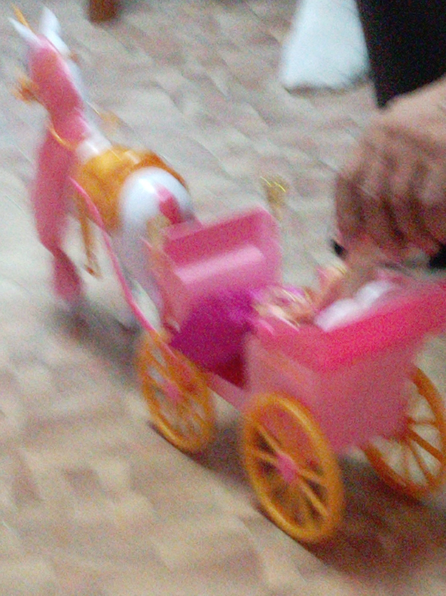 A blurred, motion-captured image shows a pink toy horse-drawn carriage. The horse, also pink with yellow accents, is facing left, its front legs extended. The carriage itself is primarily pink with yellow wheels. The background consists of a light-colored, textured surface, possibly a floor. The image quality is low, with significant motion blur, making precise identification of details difficult. There is no discernible location in the city of Artyom, Russia, or any specific environmental context provided by the image itself. The scene appears to be indoors, on a flat surface. The motion blur suggests movement, likely of the toy carriage.