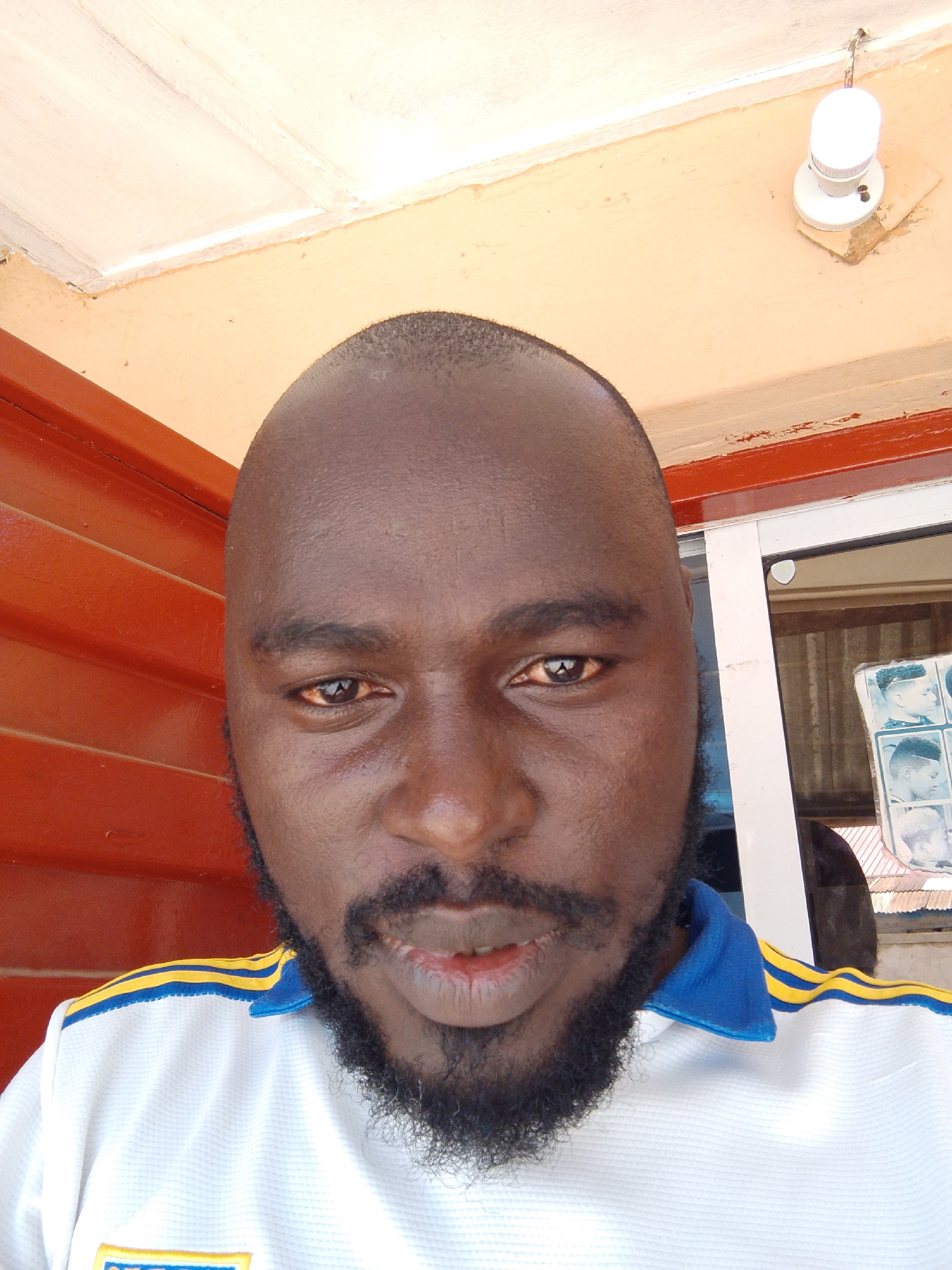 A man with dark skin, a shaved head, and a full dark beard and mustache is featured in a close-up selfie, looking directly at the camera with a neutral expression and slightly parted lips. He is wearing a white polo shirt with a blue collar and yellow stripes along the shoulders.
The background suggests an outdoor or semi-open setting, possibly a building entrance or porch in Jos, Nigeria. To the man's left, a reddish-orange wall with horizontal slats is visible. To his right, a window or glass pane reveals blurred images that appear to be posters of various hairstyles, indicating the location is likely a barbershop or hair salon. Above the man, a light peach-colored wall or beam supports a white ceiling or overhang, where a simple white light fixture with a glowing bulb is attached. Bright, natural lighting suggests it is daytime. No discernible text is clearly visible in the image.