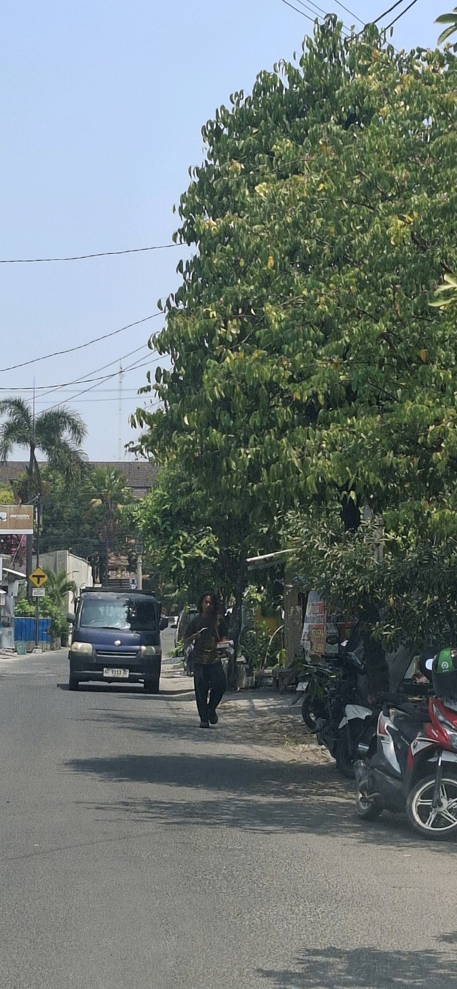 A person with long, dark hair, wearing a dark top and dark pants, walks on the sidewalk next to a road in Surakarta, Indonesia. The person is carrying a white box. A dark blue van is parked on the road to the left of the person. Various motorcycles are parked on the sidewalk to the right of the person. Several trees with green leaves are present in the scene. Buildings and power lines are visible in the background. The sky is clear and blue.