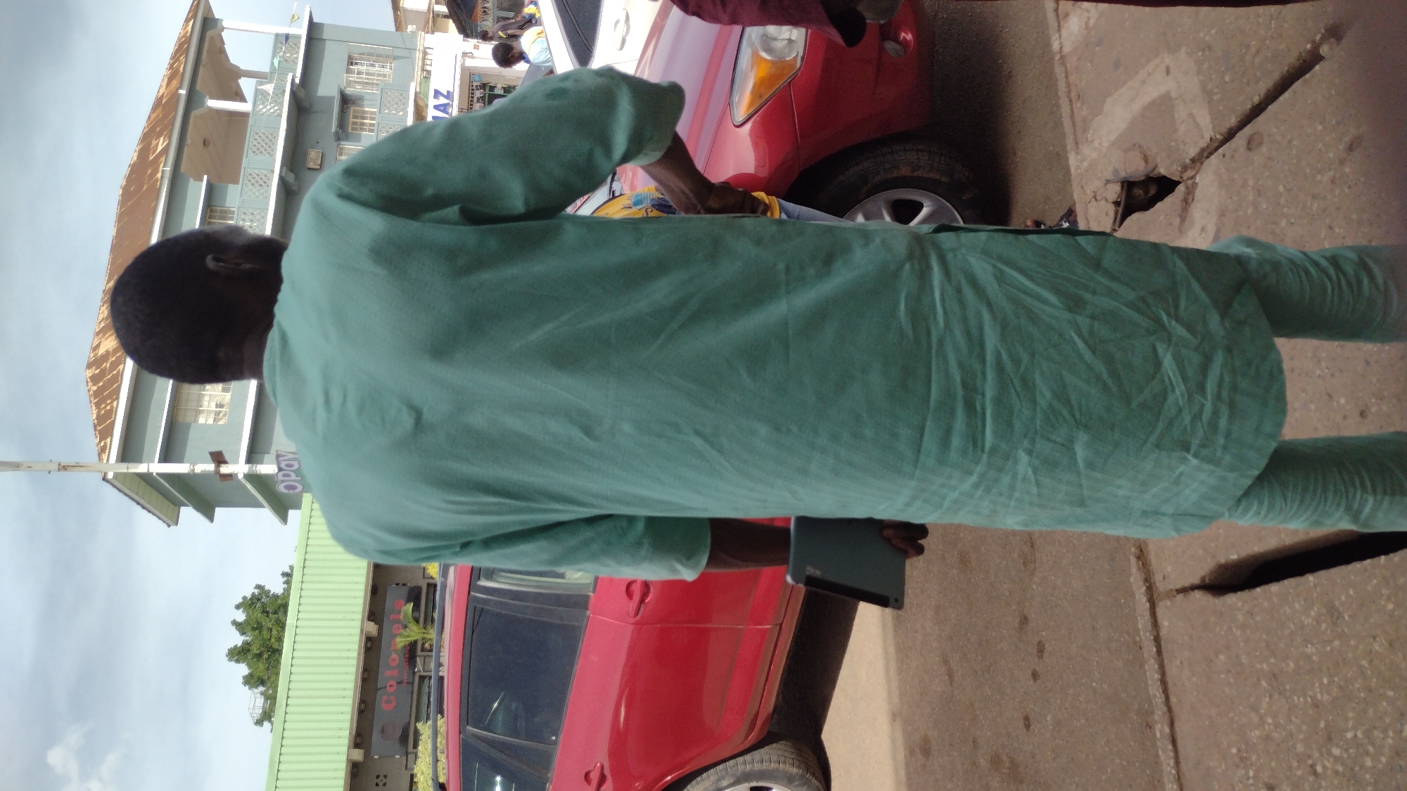 A person in a light green outfit is seen from the back, standing next to a red car. The individual is holding a tablet or similar device. The setting appears to be an urban street with a building visible in the background; it features the word "OPay" on its front. A partial view of a sign for "Colonel's" is visible. The location is likely in Jos, Nigeria, based on the request. The individual appears to be wiping the car down. The street is paved.