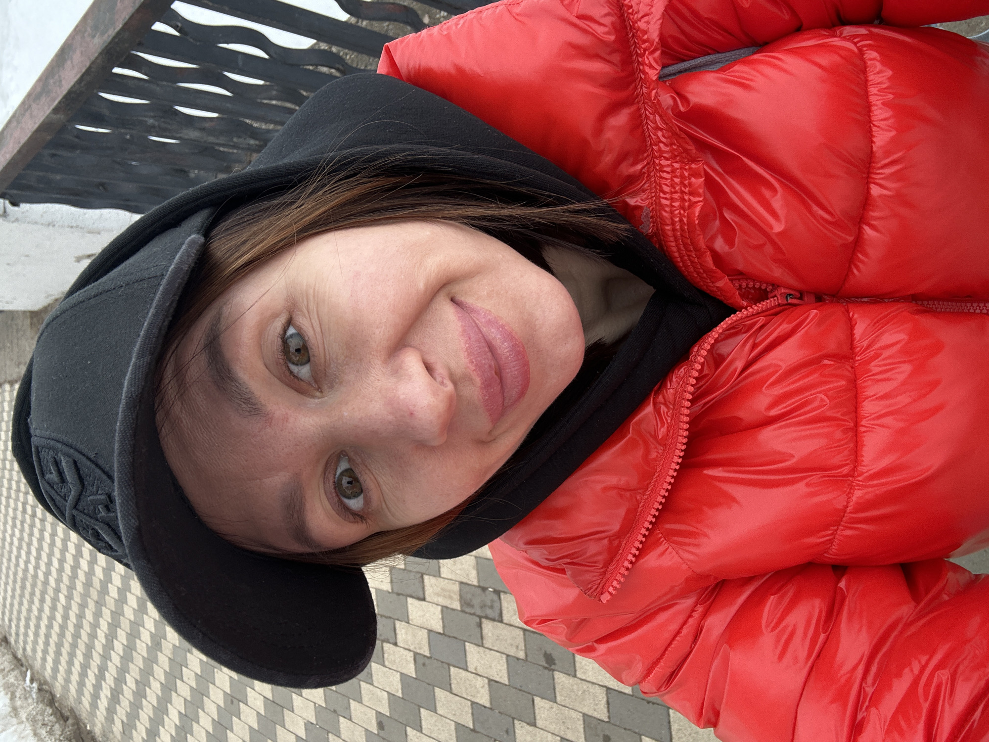 The image depicts a female individual in an outdoor setting in Votkinsk, Russia, captured in a close-up self-portrait. The subject has medium-brown hair, wide brown eyes, and a slight smile with closed lips. She wears a black baseball cap featuring an embossed emblem on the front, a black hooded sweatshirt visible beneath a bright red, glossy puffer jacket that is fully zipped. The background to the lower left shows a patterned sidewalk composed of light and dark grey rectangular paving stones arranged diagonally. To the upper right, a dark metal railing with ornate, wave-like vertical balusters is visible, behind which appears a white expanse, likely snow or ice, possibly indicating a frozen body of water or ground.