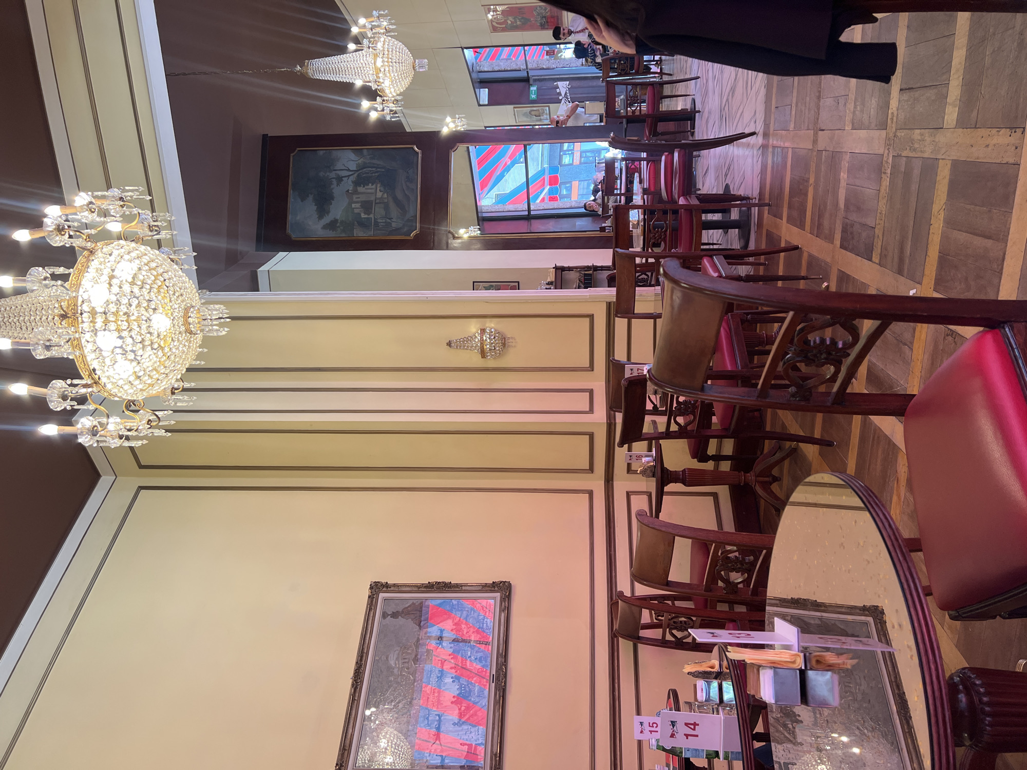 The image captures the interior of a cafe in Milan, Italy. The cafe is characterized by its classical decor, featuring crystal chandeliers, framed paintings on the walls, and dark wood furniture with red leather accents. The floor is made of wood, arranged in a geometric pattern. Sunlight streams in through a window, partially obstructed by red and white awnings, and illuminates the interior space. Tables are arranged in rows, with chairs neatly aligned, some with table numbers displayed. There are a few patrons present, one of whom is wearing a dark coat and can be seen in the upper portion of the image. The overall atmosphere is one of refined elegance.