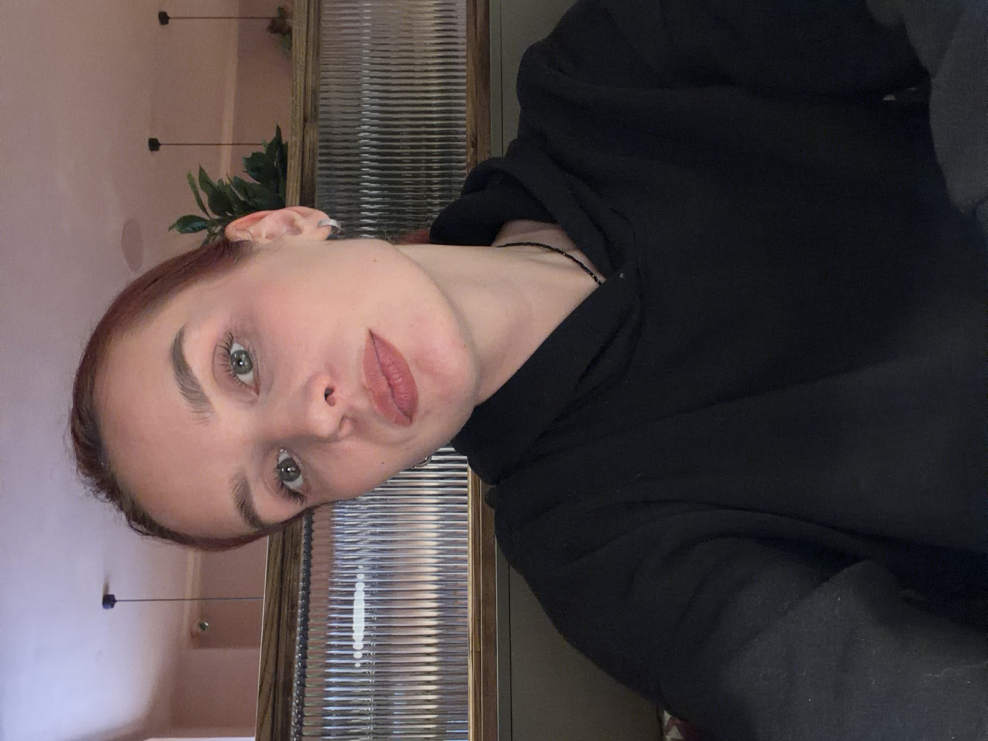 A young woman is pictured indoors. She is fair-skinned with red hair. She has light green eyes, light pink lips and defined eyebrows. She is wearing a black hoodie. A delicate black necklace is visible on her neck. The background features a wall with horizontal slats and a wooden frame. There are plants visible in the background. The wall behind her is a light pink color with visible hanging light fixtures. It is impossible to determine if the location is in the city of Chaikovsky, Russia from the image.