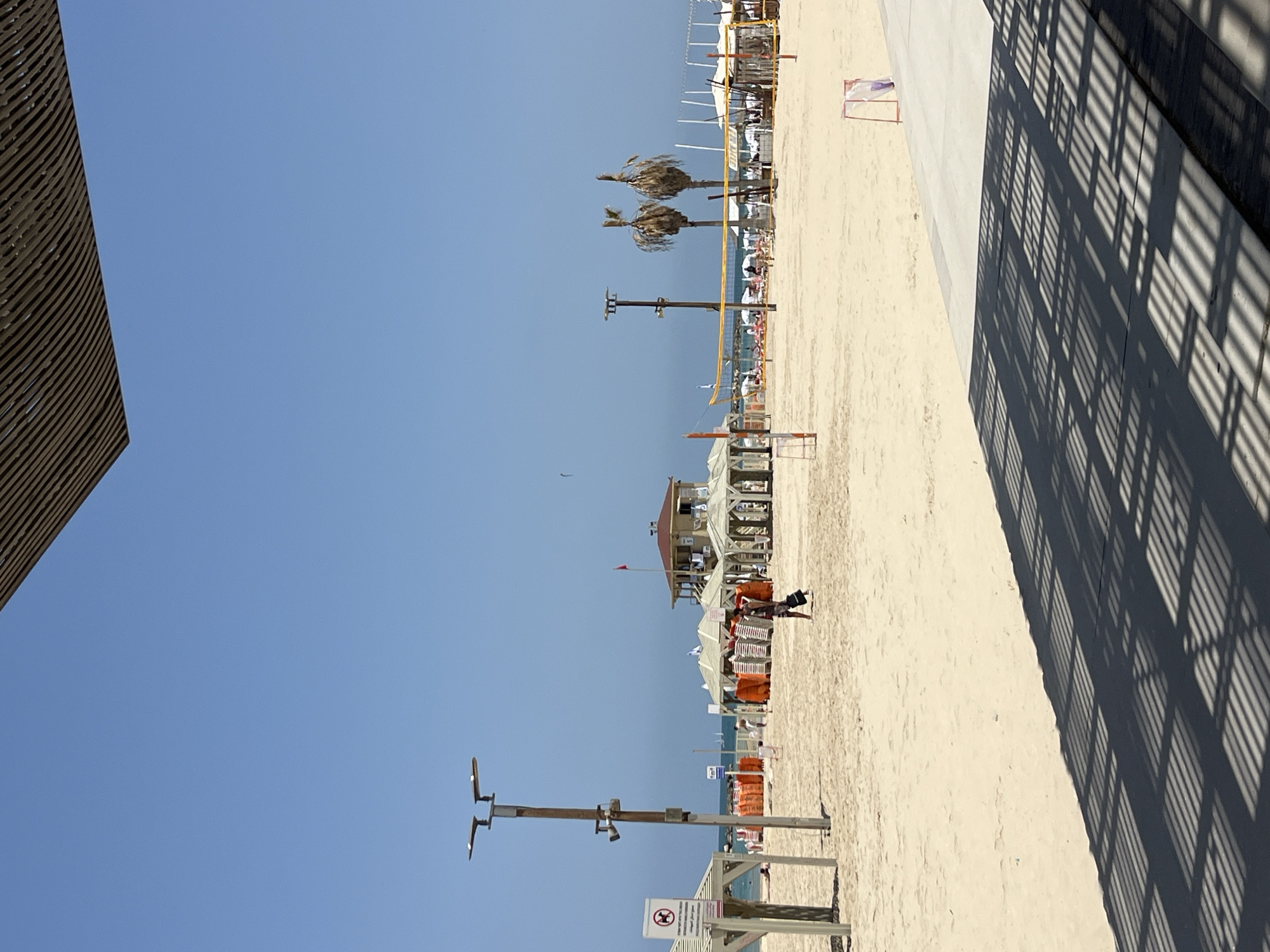 The image depicts a sunlit beach scene in Tel Aviv, Israel, under a clear blue sky. Sandy beach and a concrete walkway are visible. The shadow of a structure is cast upon the walkway, creating a patterned effect. The beach is furnished with sun umbrellas, chairs, and recreational volleyball nets. Structures, including lifeguard towers and beachside amenities, are present. People are sparsely scattered across the beach. The image is oriented vertically.