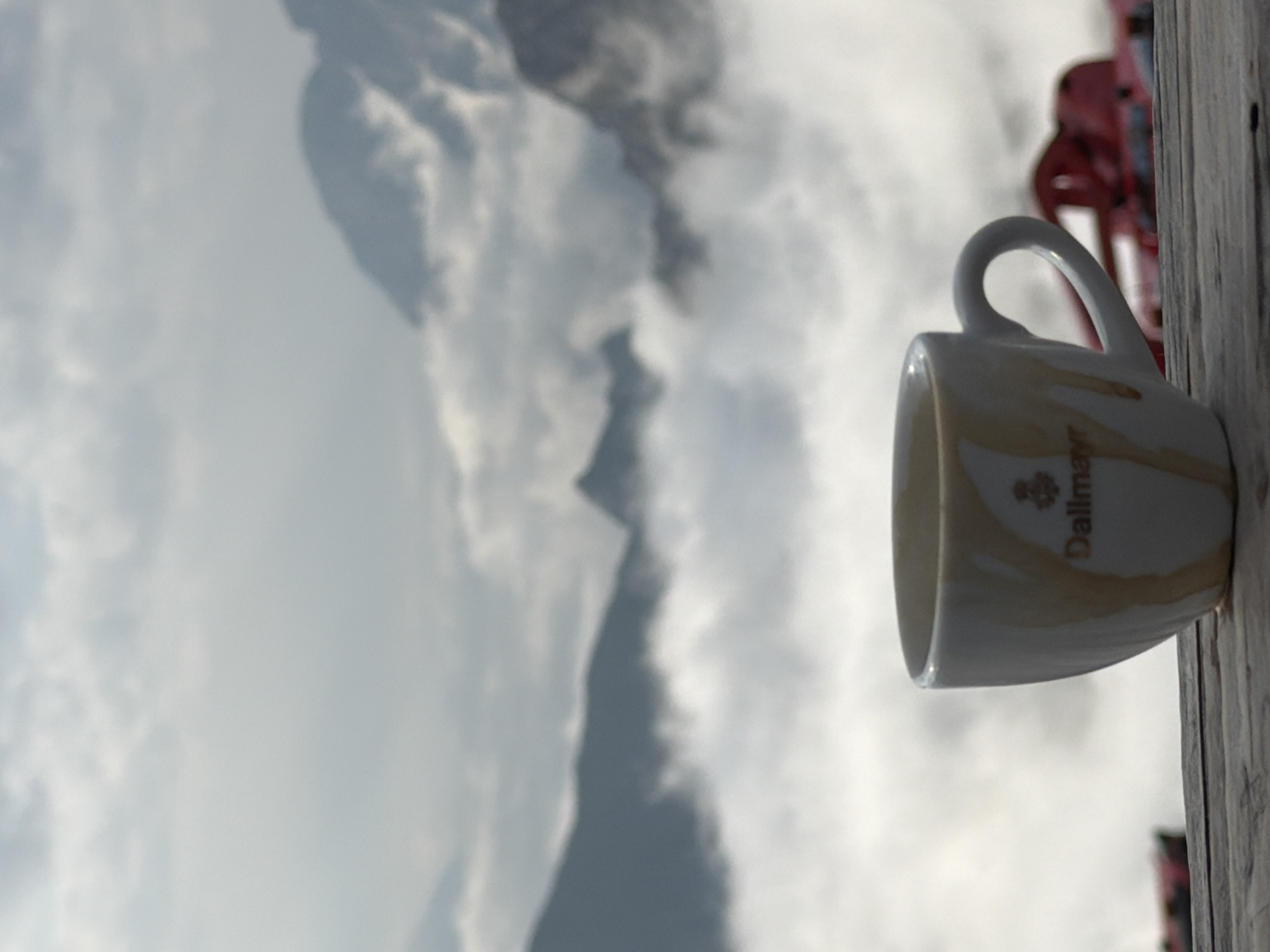 A white ceramic coffee cup with "Dallmayr" printed in gold lettering sits on a wooden table. The cup has drippings of a light brown liquid on its exterior, suggesting it held coffee. In the background, a vast expanse of clouds fills the lower half of the frame, with jagged mountain peaks emerging from the misty blanket. The sky above is overcast with diffused light. To the right of the cup, a blurry red object, possibly part of a railing or structure, is visible. The scene evokes a serene, high-altitude setting, consistent with locations like the Zugspitze in Garmisch-Partenkirchen, Germany, where one might enjoy a coffee above the clouds.
