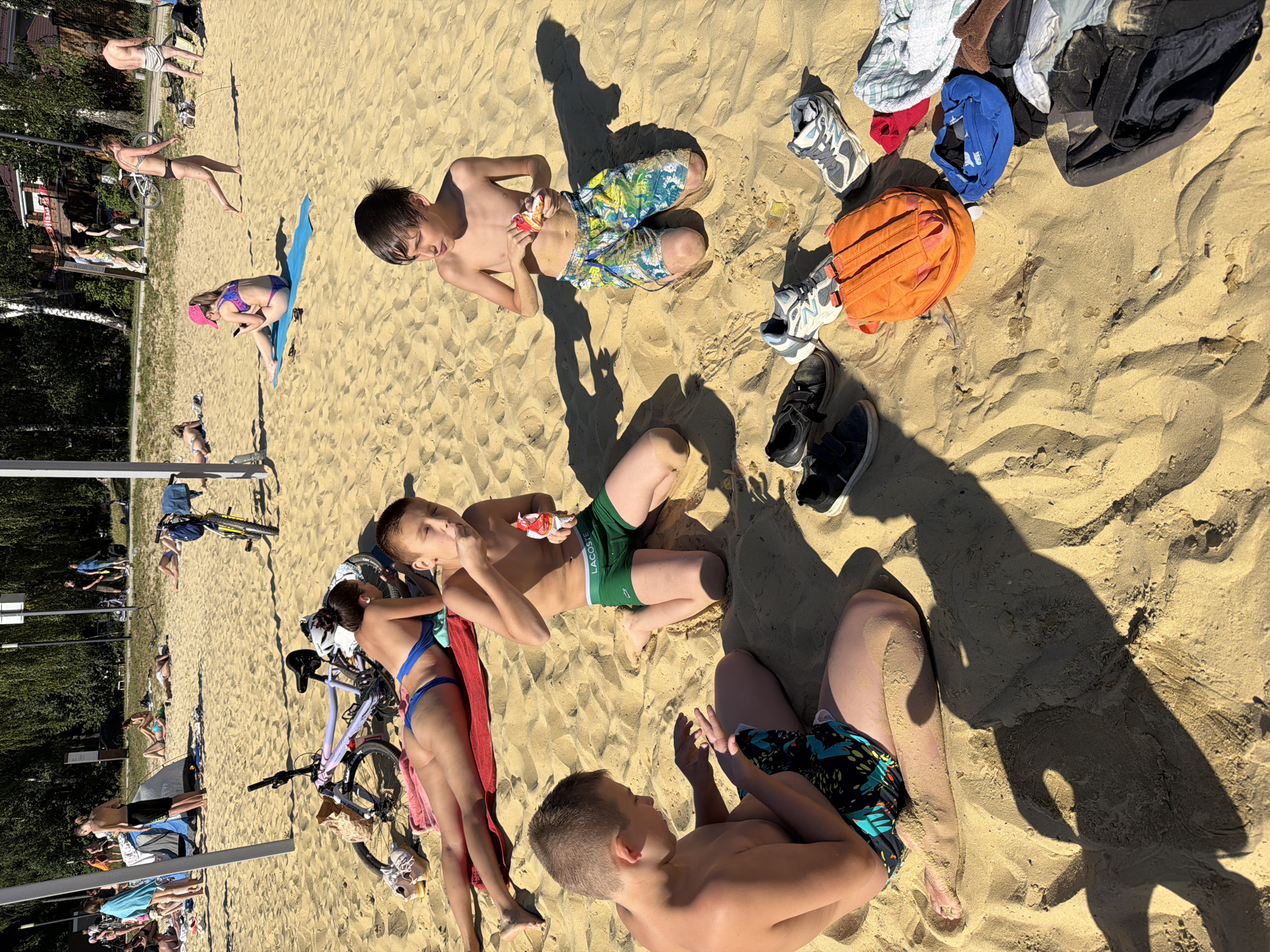 This image depicts a sunny day on a sandy beach, possibly in Malakhovka, Russia, given the provided context. In the foreground, three young boys are seated on the sand. The boy on the left is wearing patterned swim trunks and is looking down at his hands. The boy in the center is wearing green swim trunks with a "LACOSTE" logo and is eating from a small packet. The boy on the right is wearing colorful patterned swim trunks and is also eating from a packet. Scattered around the boys are several pairs of shoes, an orange backpack, and various folded clothes.
In the mid-ground, a woman in a blue bikini is lying on a red towel, facing away from the camera. A bicycle with a pink frame is parked nearby. Further back, other beachgoers are visible, some relaxing on the sand and others standing. The background shows a line of trees and some distant structures, suggesting a park or recreational area adjacent to the beach. The overall atmosphere is one of leisure and outdoor activity on a warm day.