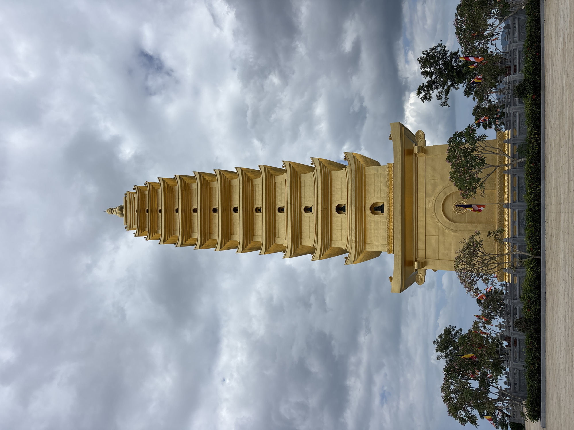 A tall, golden pagoda stands in the center of the frame, reaching towards a cloudy sky. The pagoda has a tiered structure with multiple levels, each featuring small, arched openings. At the very top, a decorative finial is visible. The pagoda is surrounded by a low, ornate stone wall and lush green bushes. Several trees with sparse foliage are planted behind the wall, and colorful flags on poles are interspersed among them. In the foreground, a paved area leads up to the pagoda. The sky is filled with dramatic, dark grey clouds, suggesting an overcast or stormy day. This scene is located in the city of Diên Khánh, Vietnam. There are no people visible in the image.