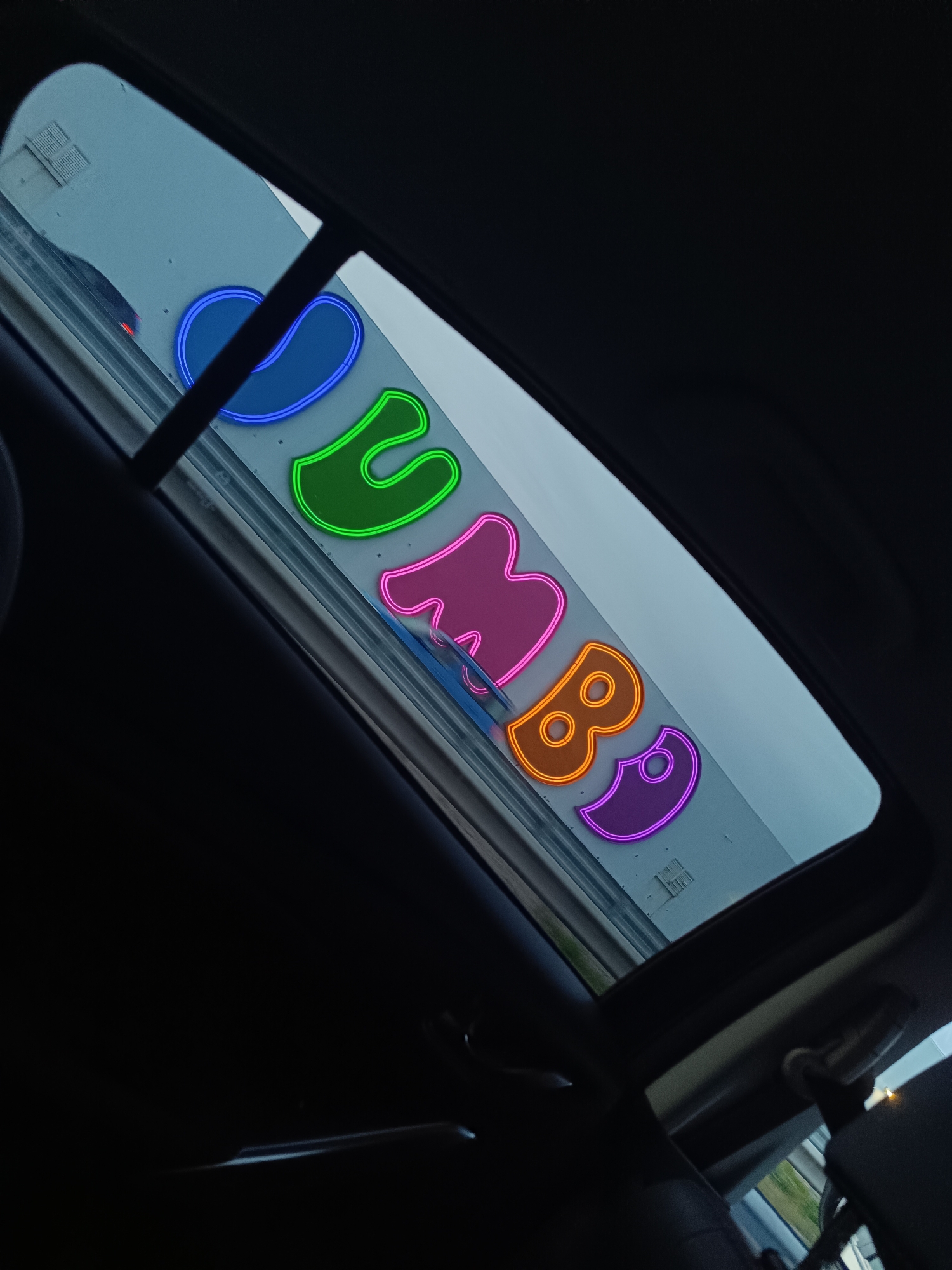 A view through the sunroof of a car shows the word "JUMBO" in neon-style lettering affixed to a building. The letters are in different colors, and the building appears to be white. The car's interior is dark, focusing attention on the exterior view.