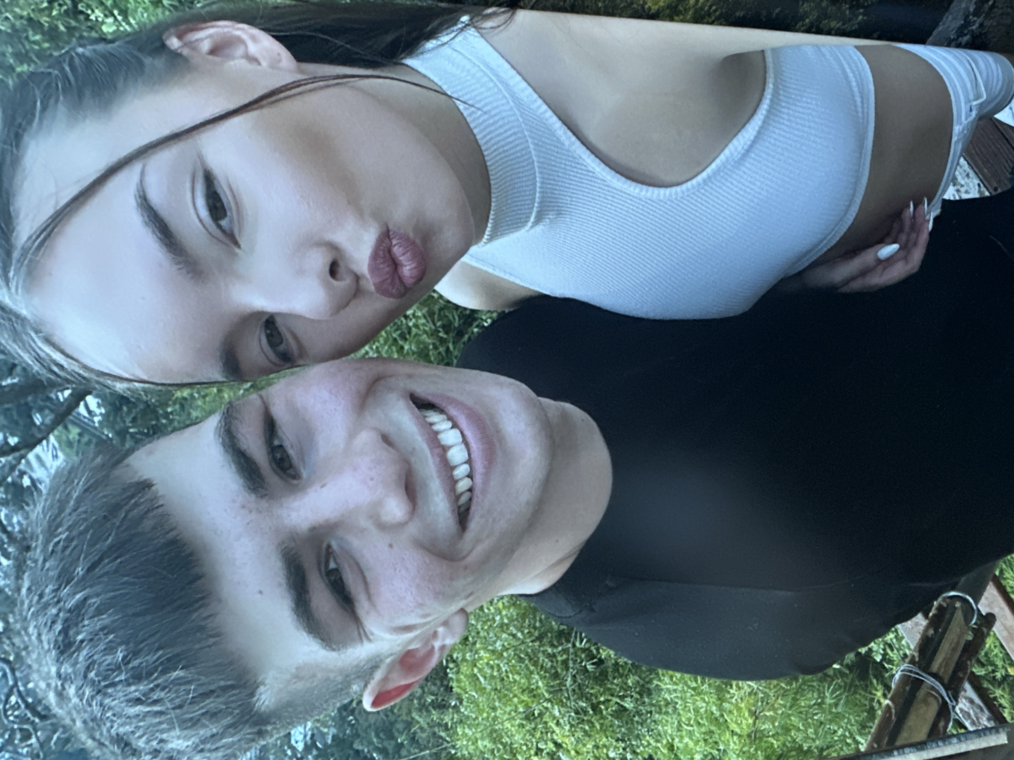 A young Caucasian couple takes a selfie outdoors. The male, on the left, has short, dark hair and is smiling broadly, showing his teeth. He is wearing a black t-shirt. The female, on the right, has long, dark hair and is puckering her lips for the camera. She is wearing a white, ribbed crop top. They are positioned in front of a lush green background of foliage. The setting appears to be a natural, possibly wooded area. There are no discernible landmarks or architectural features to indicate a specific location within Sofia, Bulgaria. The lighting suggests it is daytime.