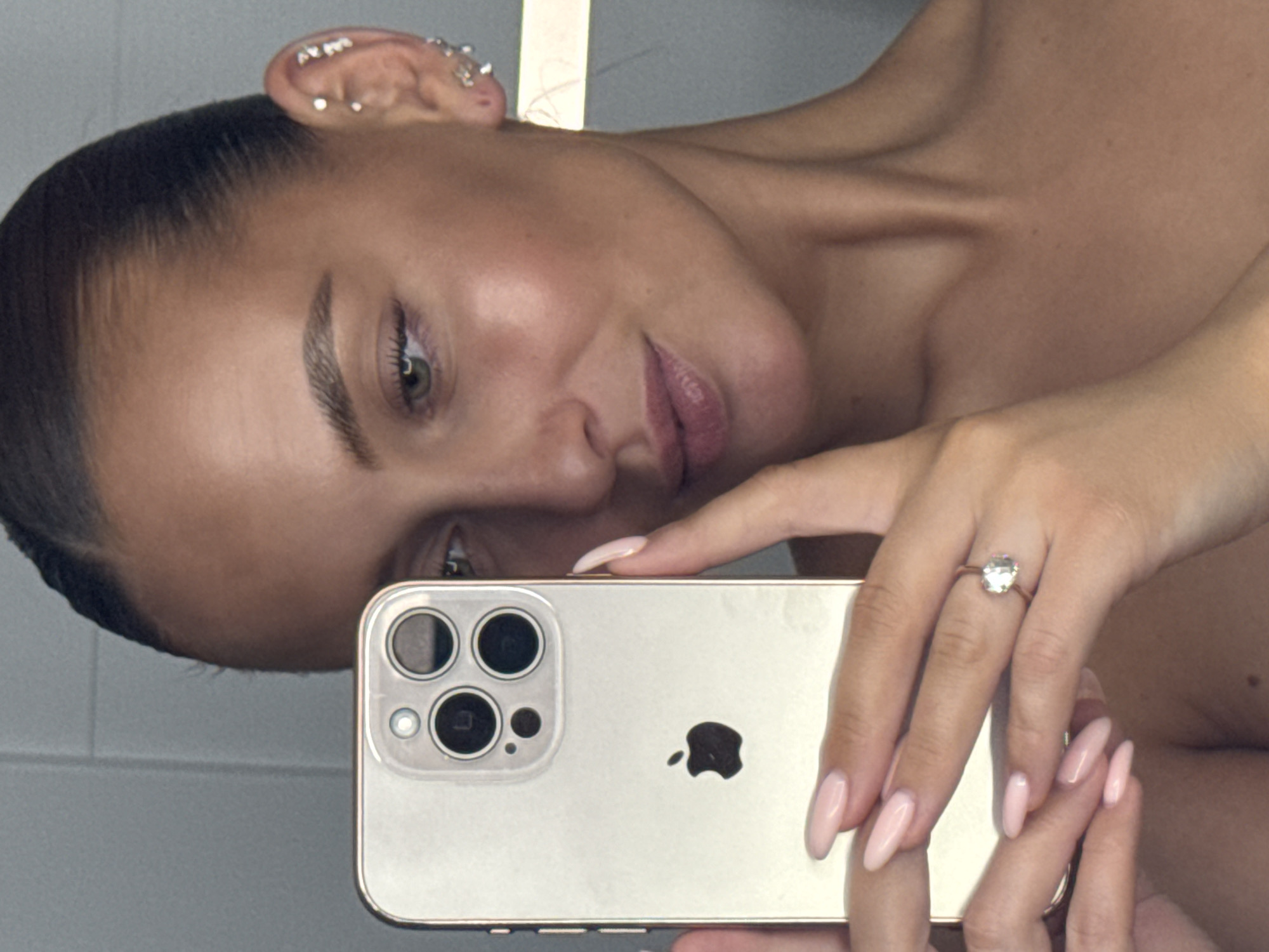 A woman takes a selfie in a mirror. She is holding a gold iPhone in her left hand, which is positioned to capture her face. Her right hand is resting on her chin, and she is wearing a diamond ring on her ring finger. She has her hair pulled back in a tight bun, and she is wearing several small earrings in her right ear. The background appears to be a bathroom with gray tiled walls. There are no discernible landmarks or details that would indicate the location is Chaikovsky, Russia.