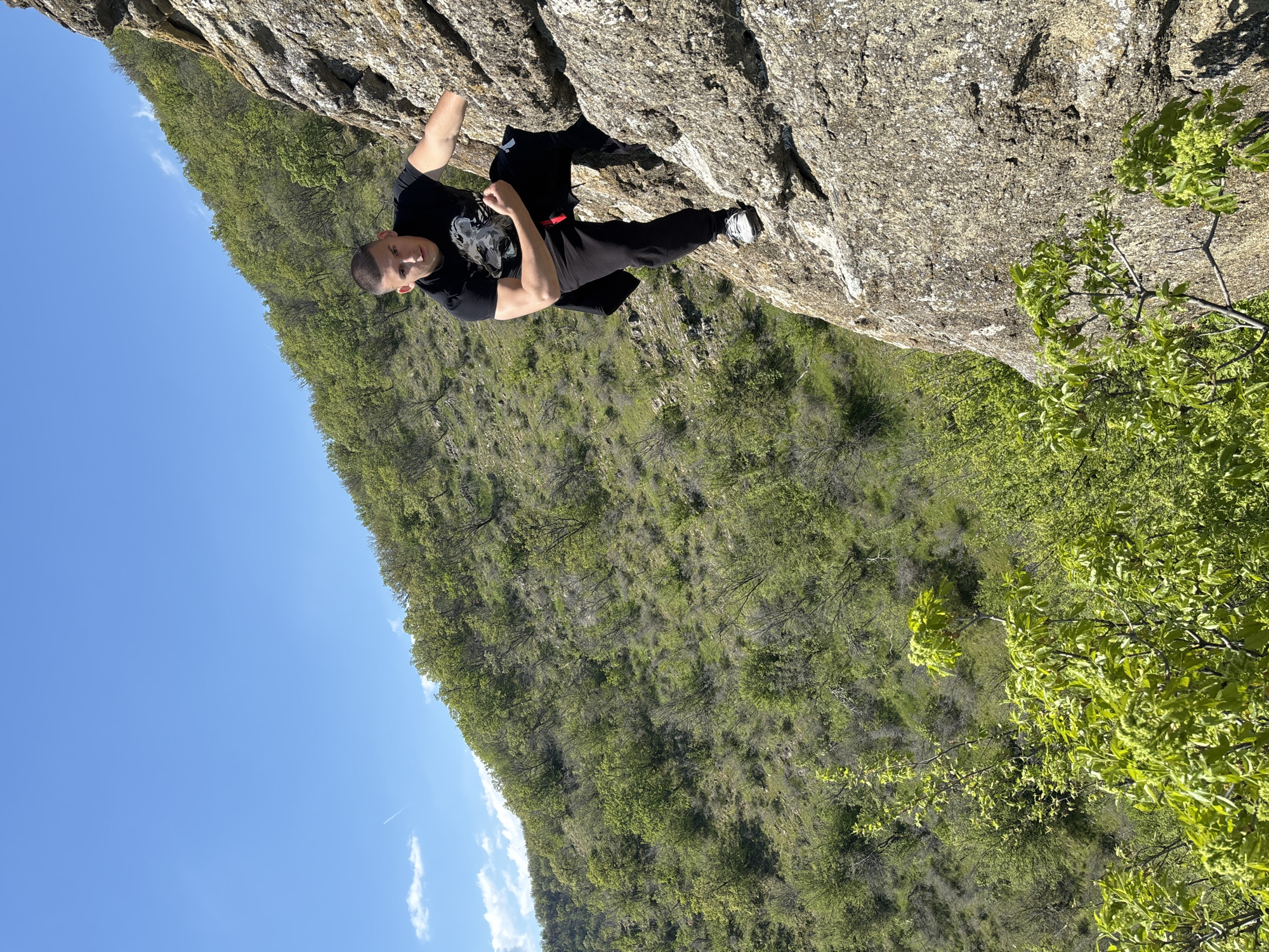 A man is rock climbing on a cliff face in Goritsa, Bulgaria. He is wearing black pants, a black t-shirt, and white sneakers. The cliff face is light brown. There are trees and other green vegetation in the background. The sky is blue with a few white clouds.