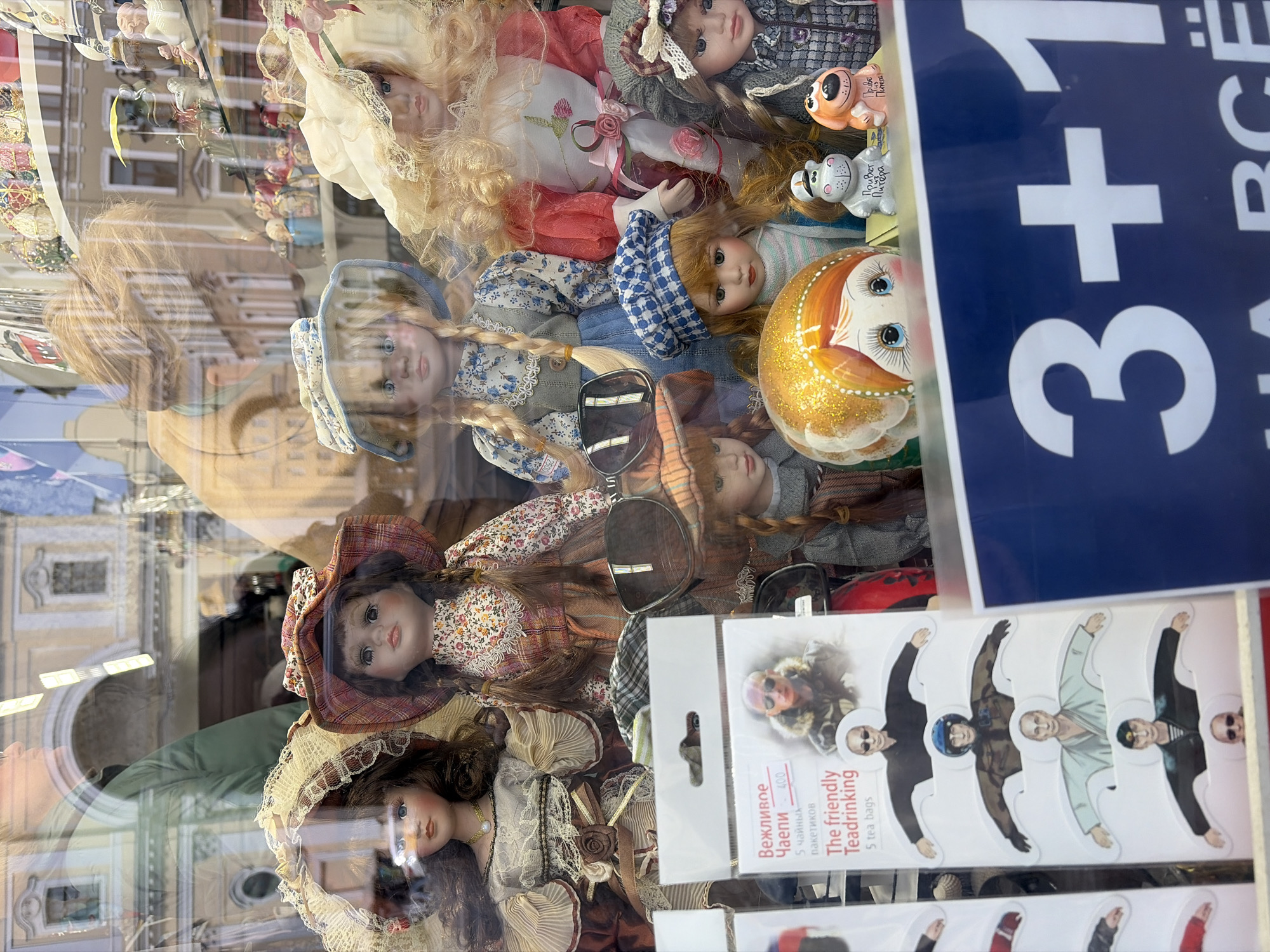 The image shows a shop window display in Saint Petersburg, Russia, filled with dolls and various souvenirs. Several porcelain dolls dressed in vintage clothing and hats are visible. Among the dolls are matryoshka dolls and other figurines. There is also a package of "The Friendly Teadrinking" tea bags featuring images reminiscent of political figures, packaged in 5 tea bags. A sign displaying "3+1" is also visible. The display is set against the backdrop of Saint Petersburg architecture, reflected in the shop window.