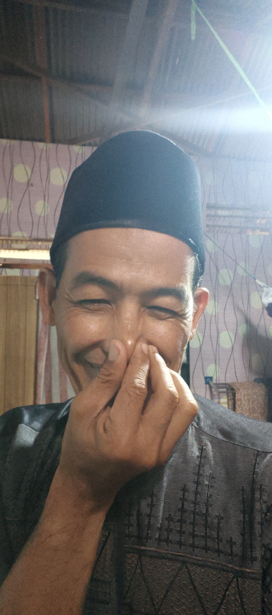 A man wearing a black peci hat and a dark gray shirt with embroidered patterns is shown from the chest up. He is holding his right hand up to his nose, with his fingers covering it. His eyes are closed, and he appears to be smiling or laughing. The background consists of a patterned wallpaper and a ceiling. The image was taken in Muaro Jambi, Indonesia.