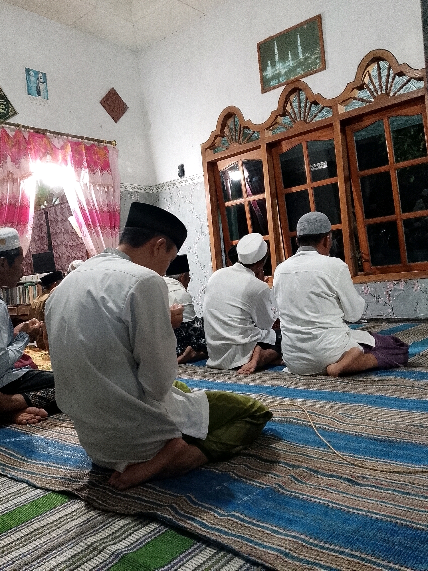 In Rejoyoso, Indonesia, a group of men are gathered indoors for prayer, seated on a striped mat on the floor. The room appears to be a private residence, with white walls and floral-patterned wallpaper near the ceiling. A large wooden window with decorative carvings spans a significant portion of the back wall, reflecting the dim interior lighting and the dark exterior. In front of the window, three men in white attire and skullcaps are positioned facing away from the viewer, with their heads bowed. To the left, a man in a white shirt and black cap, also facing away, is prominent in the foreground, with his legs crossed and a green cloth tucked beneath him. Further to the left, another man in a white shirt and light-colored cap is partially visible, also facing towards the prayer direction. The lighting in the room is dim and warm, with sunlight filtering through pink curtains on a window to the left, casting long shadows. The overall atmosphere suggests a moment of quiet devotion.