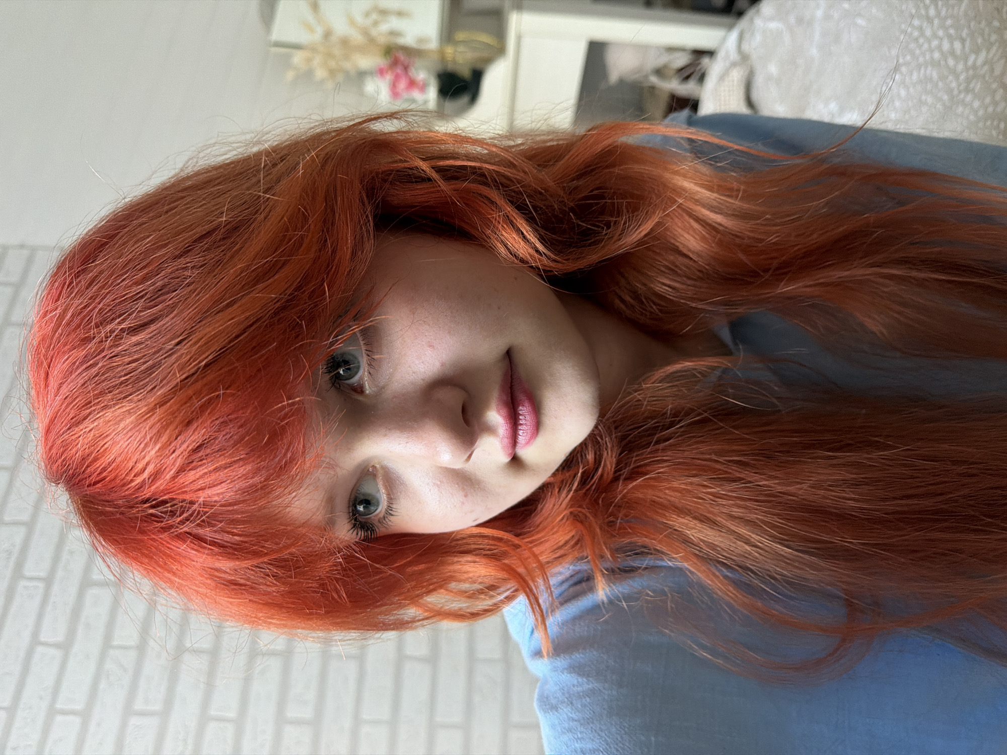 The image is a close-up, low-angle portrait of a young person with vibrant, reddish-orange hair. Their head is tilted to the side, and their gaze is directed towards the camera, with a neutral expression. The person has fair skin with freckles, blue eyes, and full lips. They are wearing a light blue garment, possibly a shirt or blouse.
The background is softly blurred, but hints of an indoor setting are visible. To the left, a white brick-patterned wall suggests an interior wall. In the upper right, a blurred, textured surface, possibly a couch or cushion, and hints of other furniture indicate a domestic environment. The lighting appears to be natural, suggesting daytime.
The image is taken from a low angle, making the subject's hair appear to cascade around their face and shoulders. The focus is sharp on the subject's face and hair, with the background rendered out of focus to emphasize the portrait. There is no visible text in the image. The setting appears to be a home environment in Sochi, Russia.